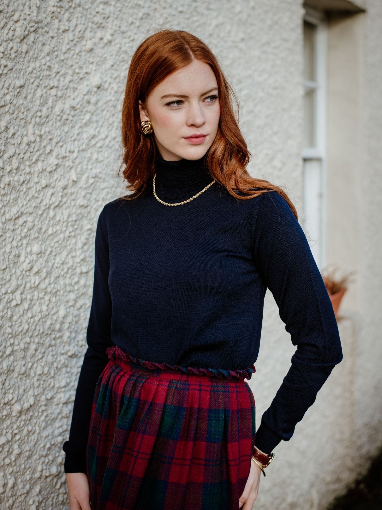 A woman with long red hair wears the Campbells of Beauly Merino Poloneck, gold jewelry, and a red and green plaid skirt as she stands against a textured white wall, looking to the side—a stylish layered look for any season.