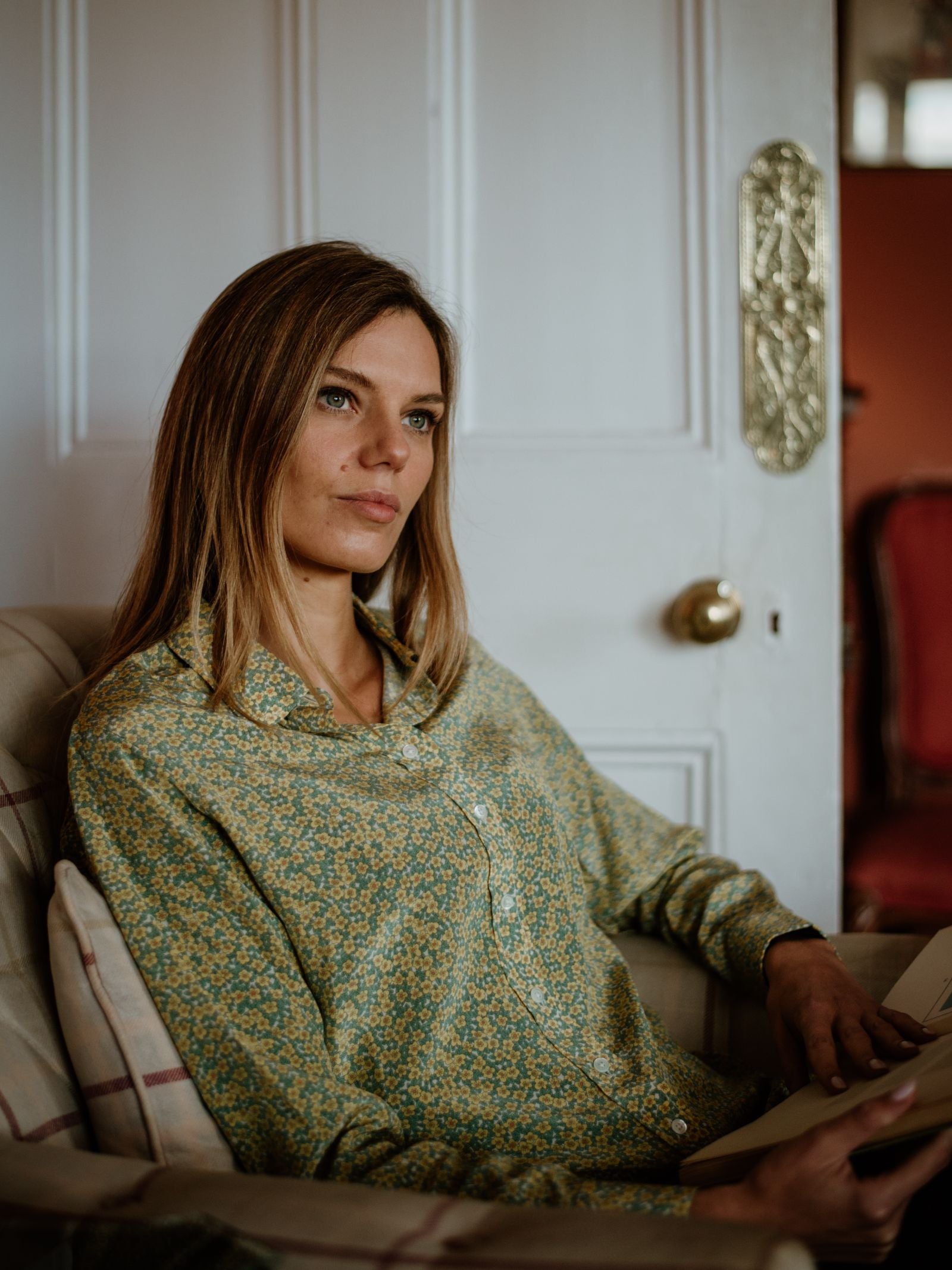 A woman with long light brown hair wears the Campbells of Beauly Liberty Print Silk Shirt, made in Portugal, as she sits thoughtfully holding a notebook; a white door and red chair are visible in the background.