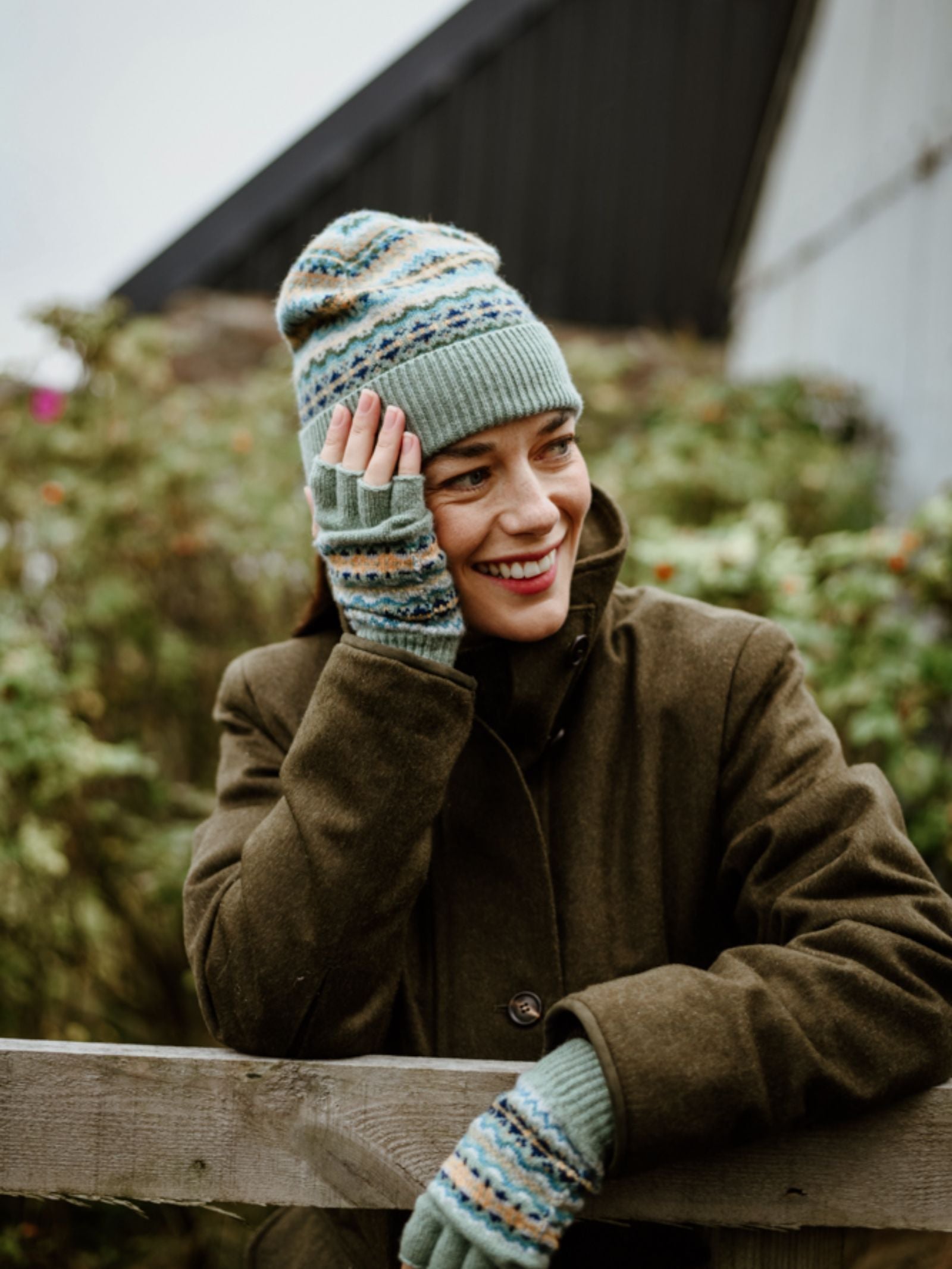 A smiling woman in a green coat stands outdoors, wearing Campbell's of Beauly Lambswool Fairisle Hat and matching fingerless gloves with blue, green, and beige patterns. She rests her hand on her face by a wooden fence with greenery behind her.