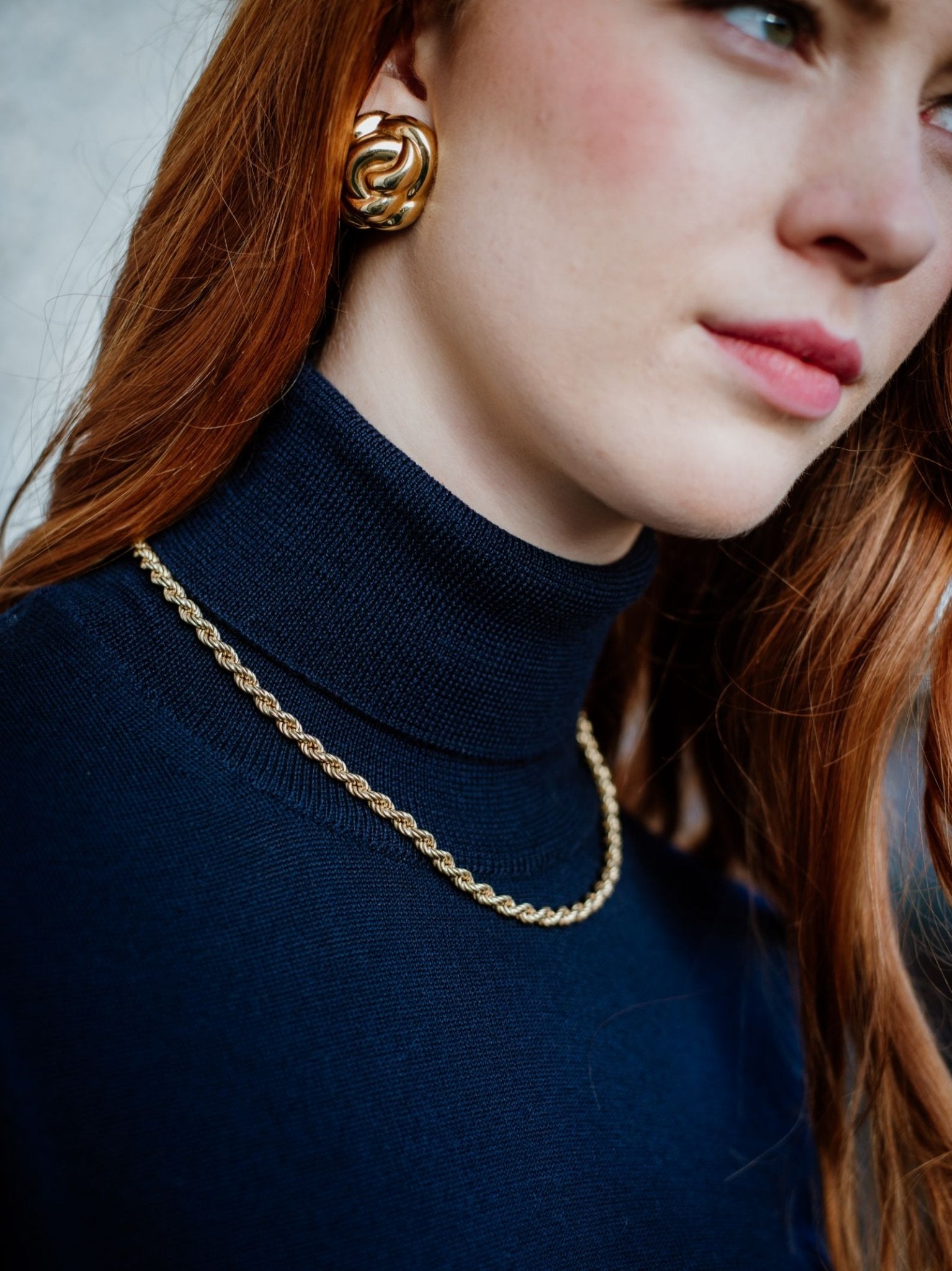 A woman with long red hair wears the Campbells of Beauly Merino Poloneck in navy, paired with gold jewelry. The cropped photo highlights the superfine merino wool texture, her necklace, and large knot earring.