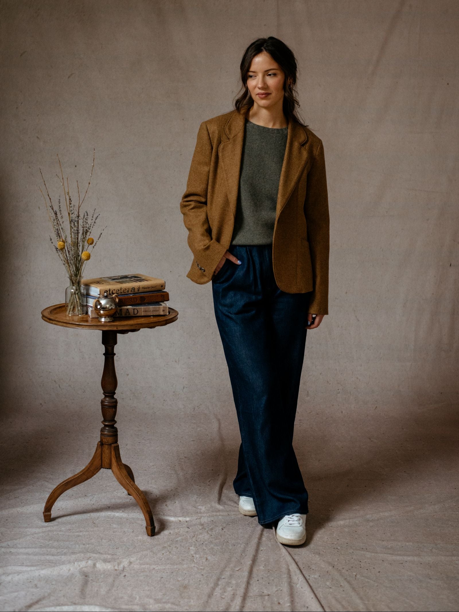 A woman stands indoors wearing the House Jacket by Campbells of Beauly over a gray sweater, wide-leg blue trousers, and white sneakers. She is beside a small round table with books, dried flowers in a vase, and a silver item against beige fabric.