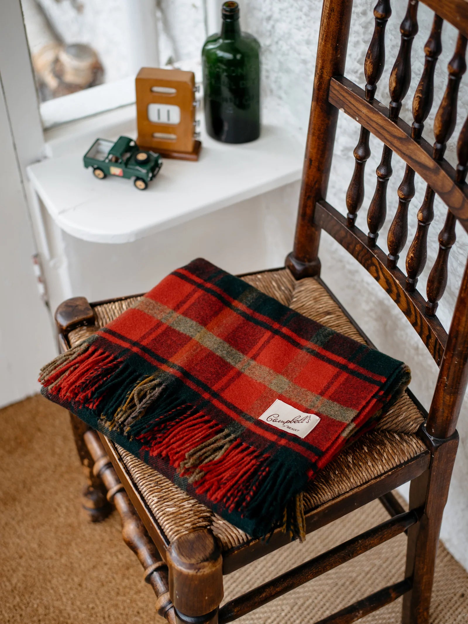 A Campbell's of Beauly Lambswool Tartan Blanket is folded on a wooden spindle-back chair. On the white windowsill behind, a toy truck, green bottle, and small wooden clock complete the cozy scene.