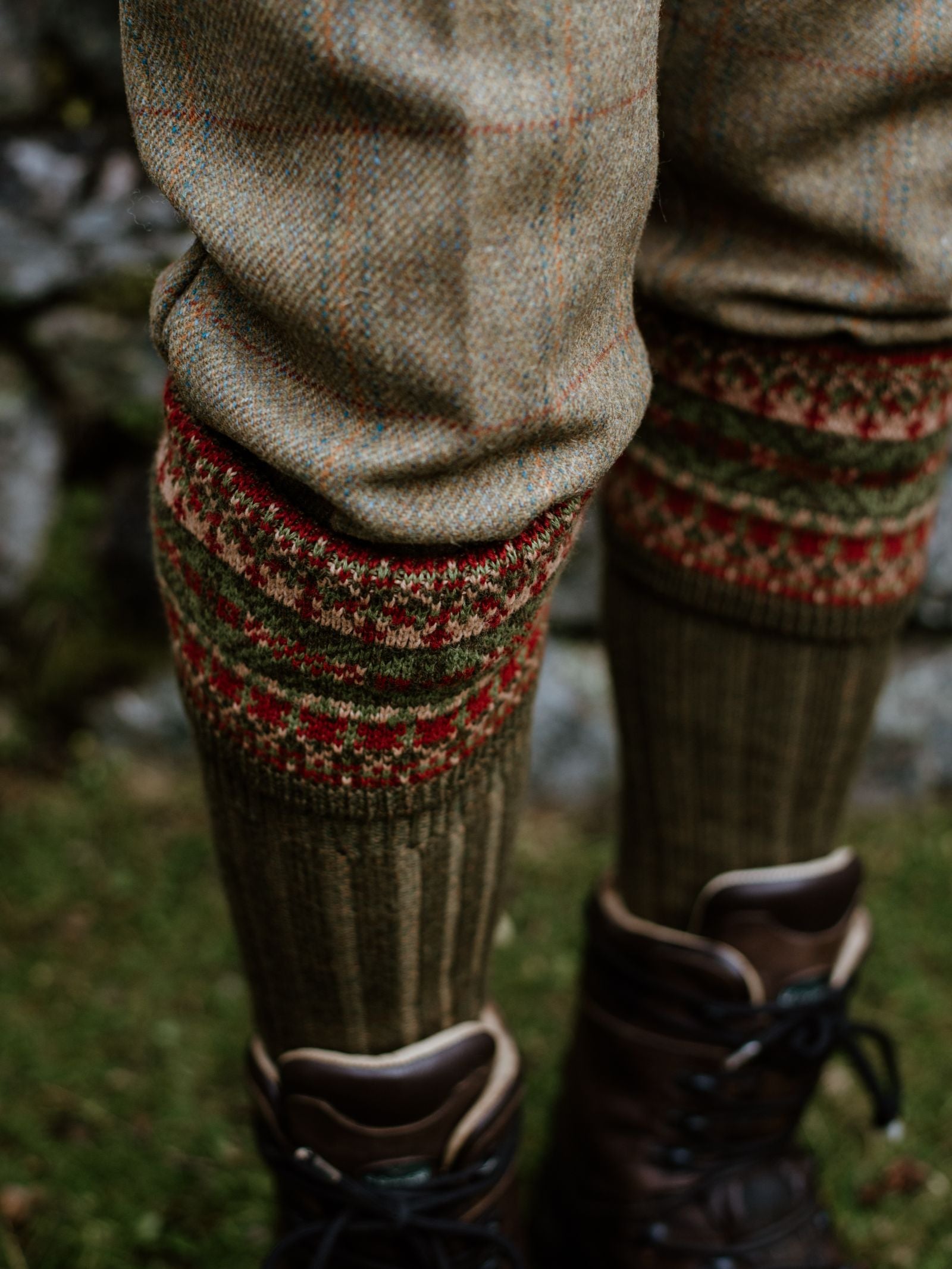 A close-up of lower legs on grass near a stone wall, wearing brown hiking boots, beige plaid pants, and Campbells of Beauly Fairisle Shooting Socks—classic knitwear heritage in a merino blend.