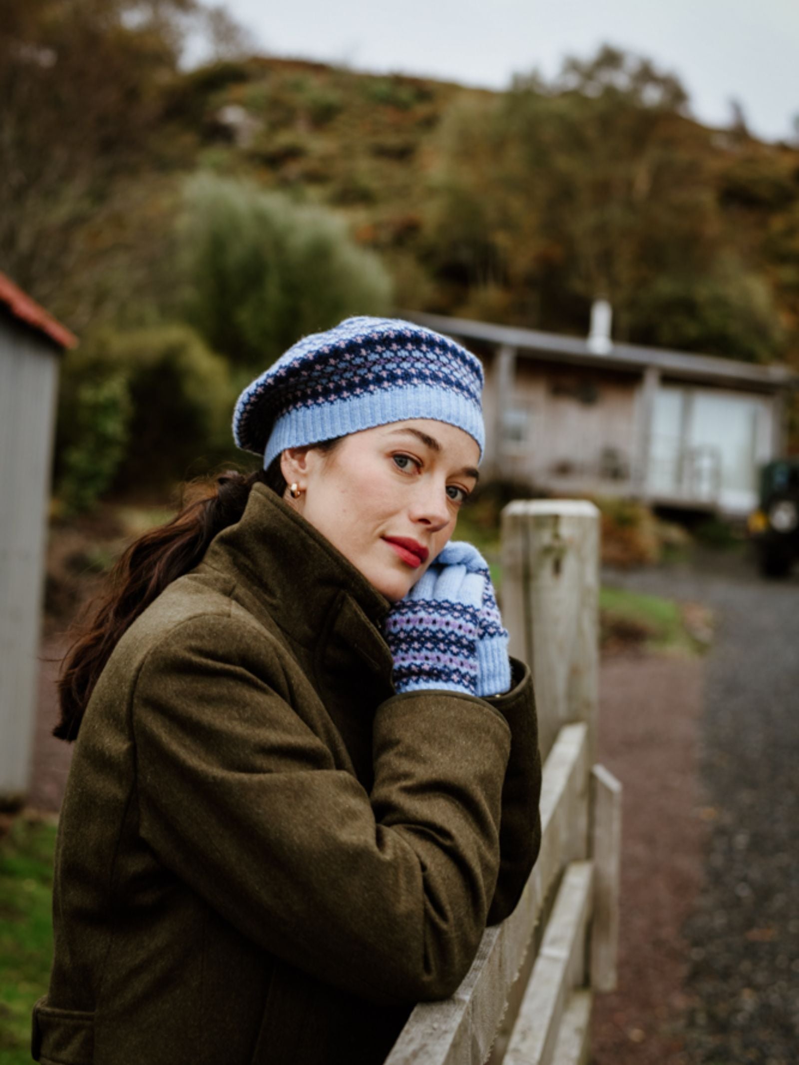 A woman in a dark coat and matching gloves wears the Campbell's of Beauly Fairisle Beret, leaning on a wooden fence with a cottage and trees in the background. She looks thoughtfully at the camera.