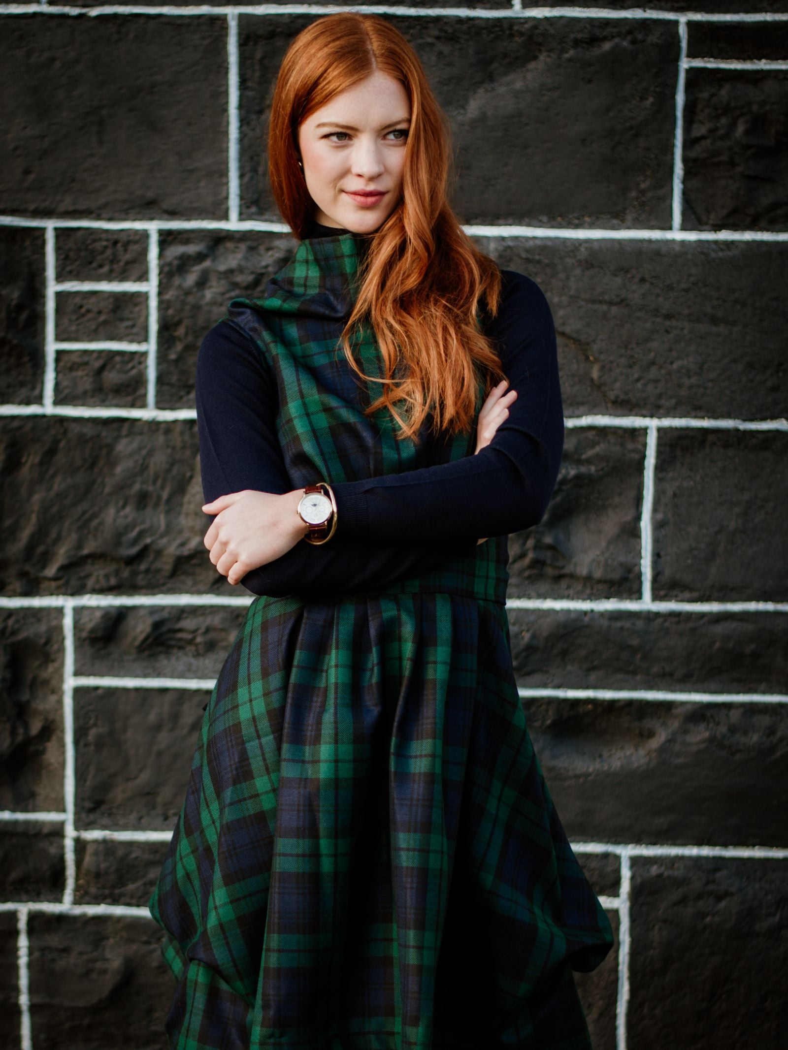 A woman with long red hair stands in front of a black brick wall, wearing a Campbells of Beauly Reversible Wool Wrap Dress over a black long-sleeve top, arms crossed and smiling slightly.