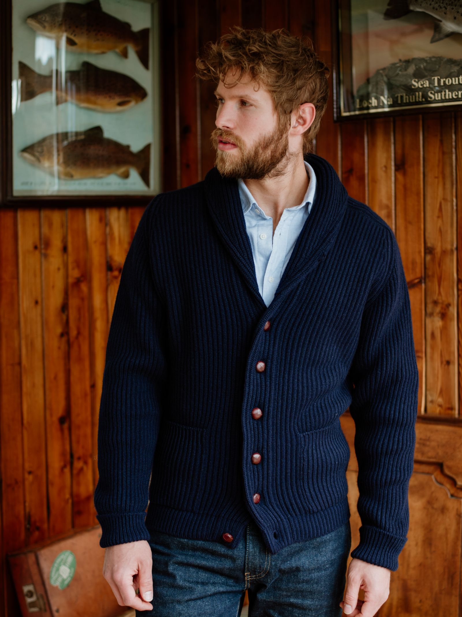 A bearded man in a navy blue Campbells of Beauly Cashmere Shawl Neck Collar Cardigan and light blue shirt stands indoors against wood-paneled walls with framed fish illustrations, looking to his left.