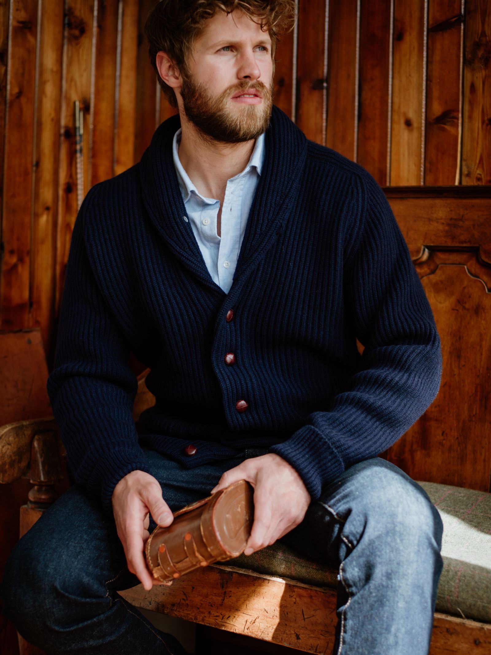 A bearded man in a Campbells of Beauly Cashmere Shawl Neck Collar Cardigan and jeans sits on a wooden bench, holding a large chocolate bar, looking off to the side as sunlight streams through the wooden backdrop.