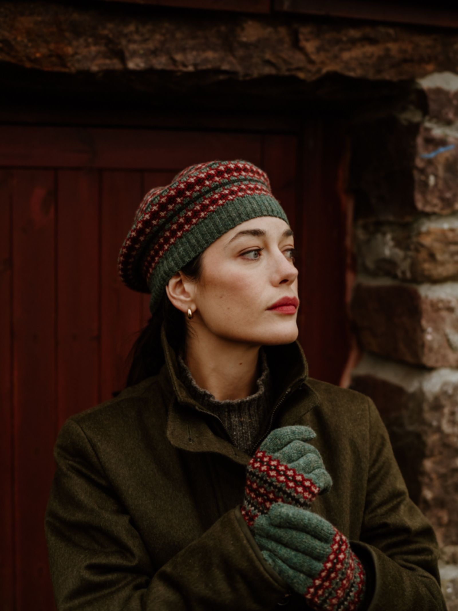 A woman in a green coat, knit gloves, and the Campbell's of Beauly Fairisle Beret stands before a rustic wooden door and stone wall, gazing thoughtfully to the side.