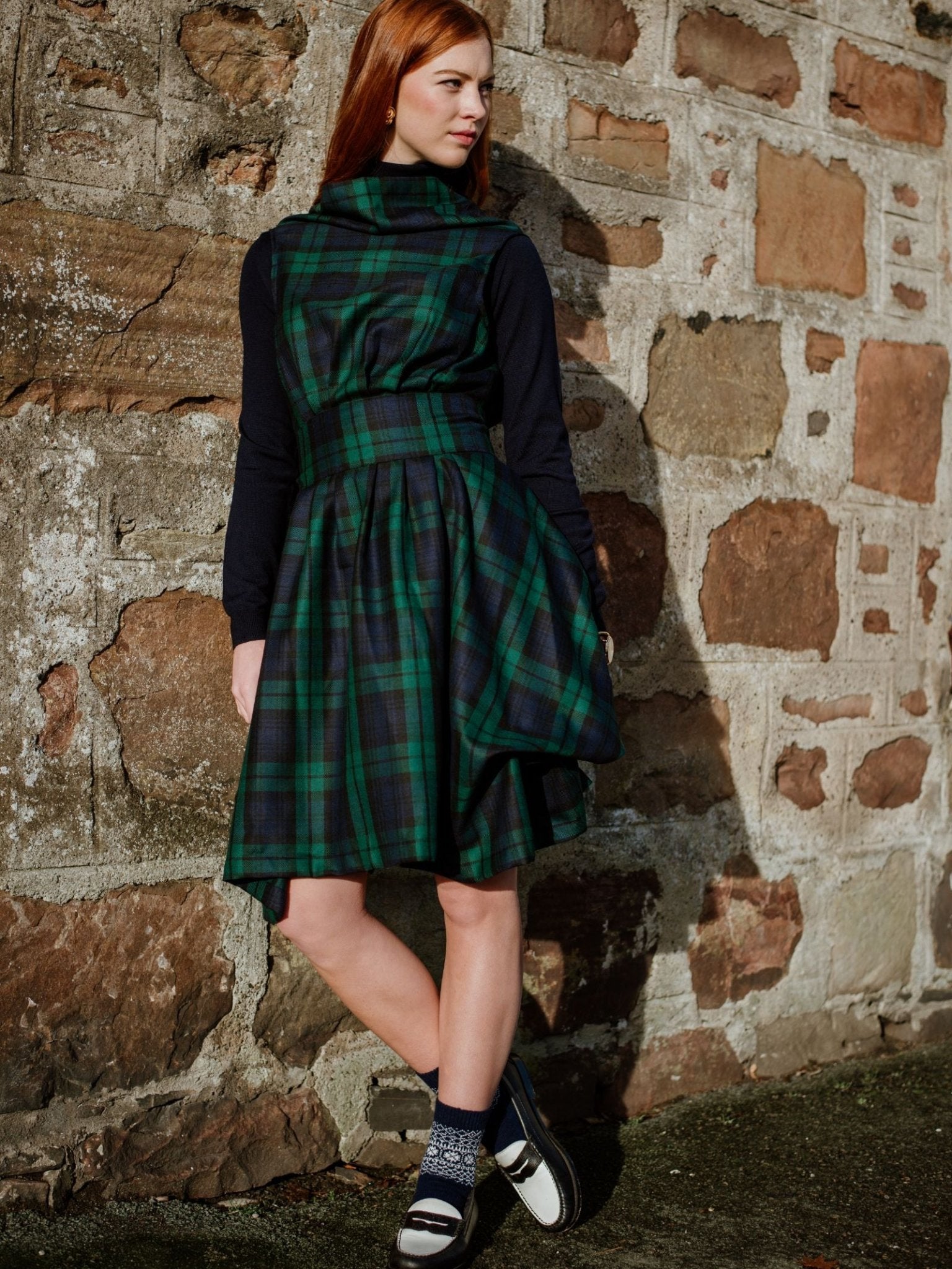A woman with long red hair stands by a stone wall in a green and navy tartan dress over a black top, white shoes with black straps, and Campbells of Beauly Two-Colour Fairisle Cashmere Socks. She looks to the side.
