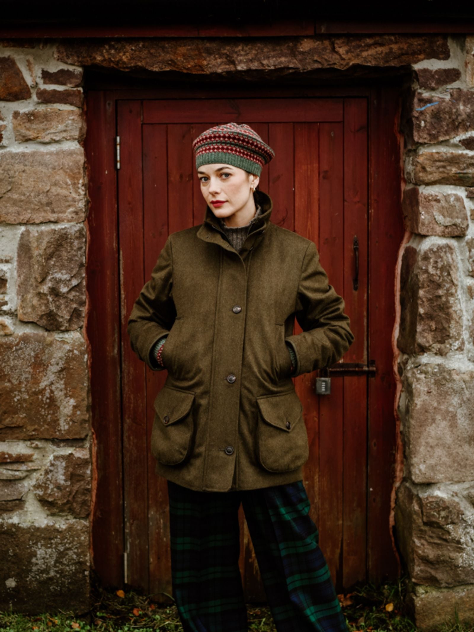 A person wearing a green coat, plaid pants, and a Campbell's of Beauly Fairisle Beret stands in front of a rustic red wooden door framed by stone walls, looking at the camera with hands in pockets.