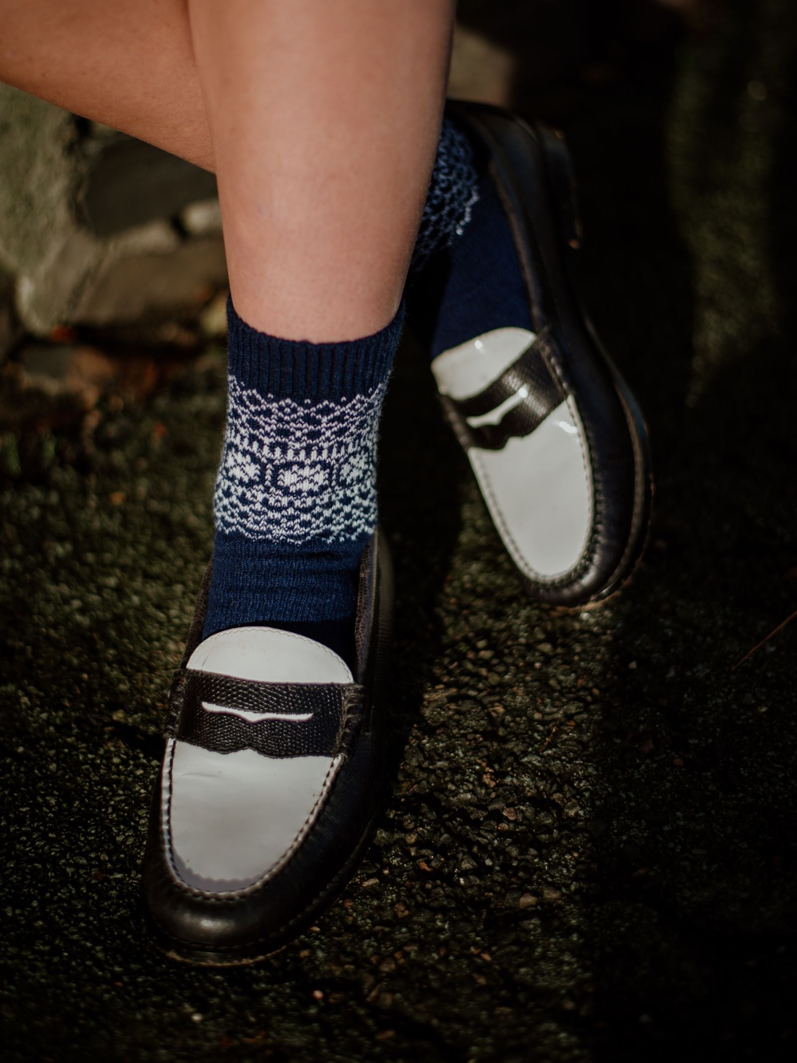 A person wearing Campbells of Beauly Two-Colour Fairisle Cashmere Socks in navy with a white pattern and black-and-white loafers stands on a dark, textured surface as sunlight casts shadows on their legs and feet.