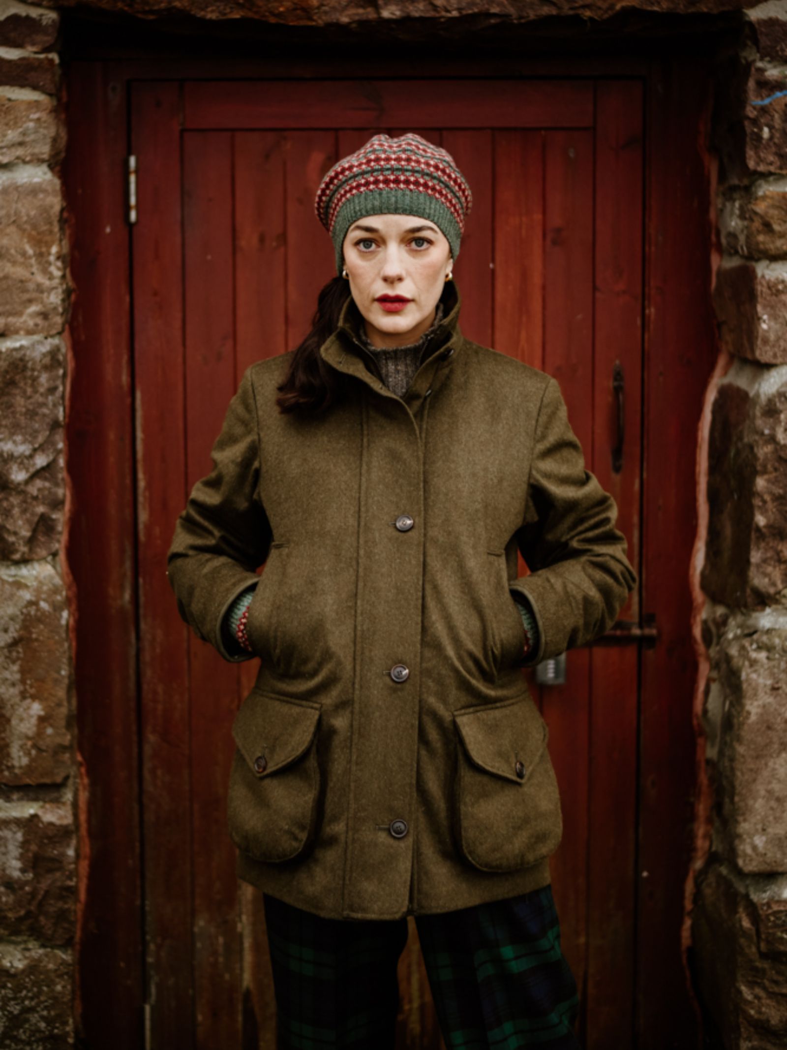A woman wearing the Campbell's of Beauly Fairisle Beret stands with hands in her pockets before a rustic red wooden door set in a stone wall, gazing seriously at the camera.