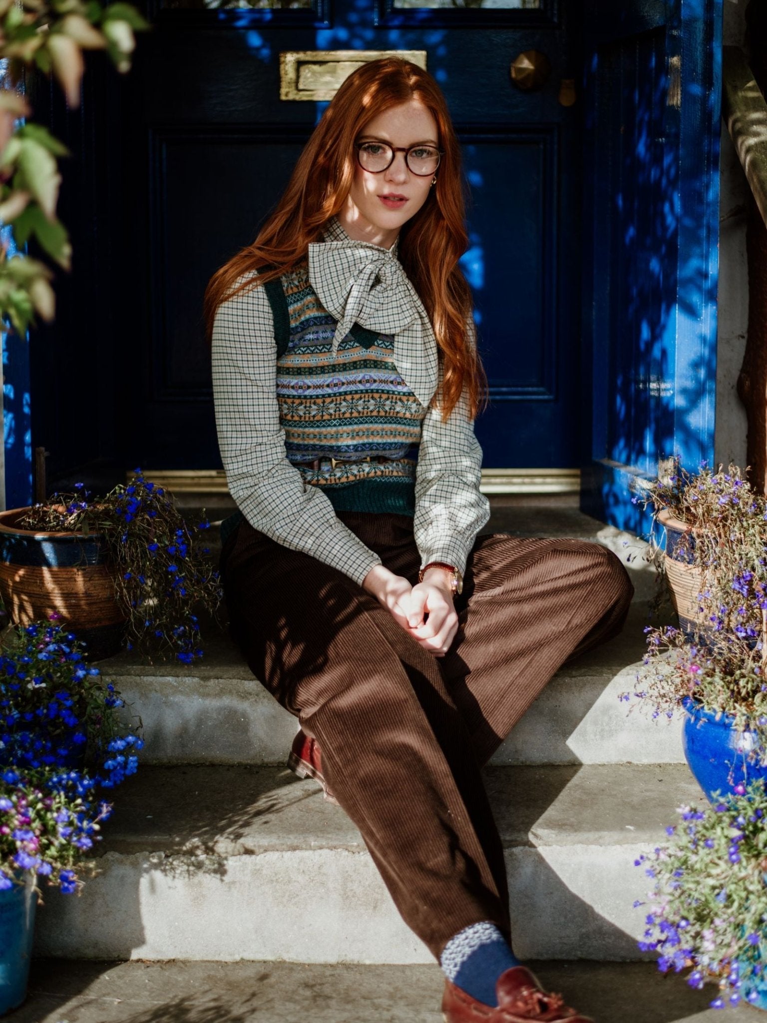 A woman with long red hair and glasses sits on stone steps by a blue door, wearing Campbells of Beauly Two-Colour Fairisle Cashmere Socks, surrounded by potted blue and purple flowers.