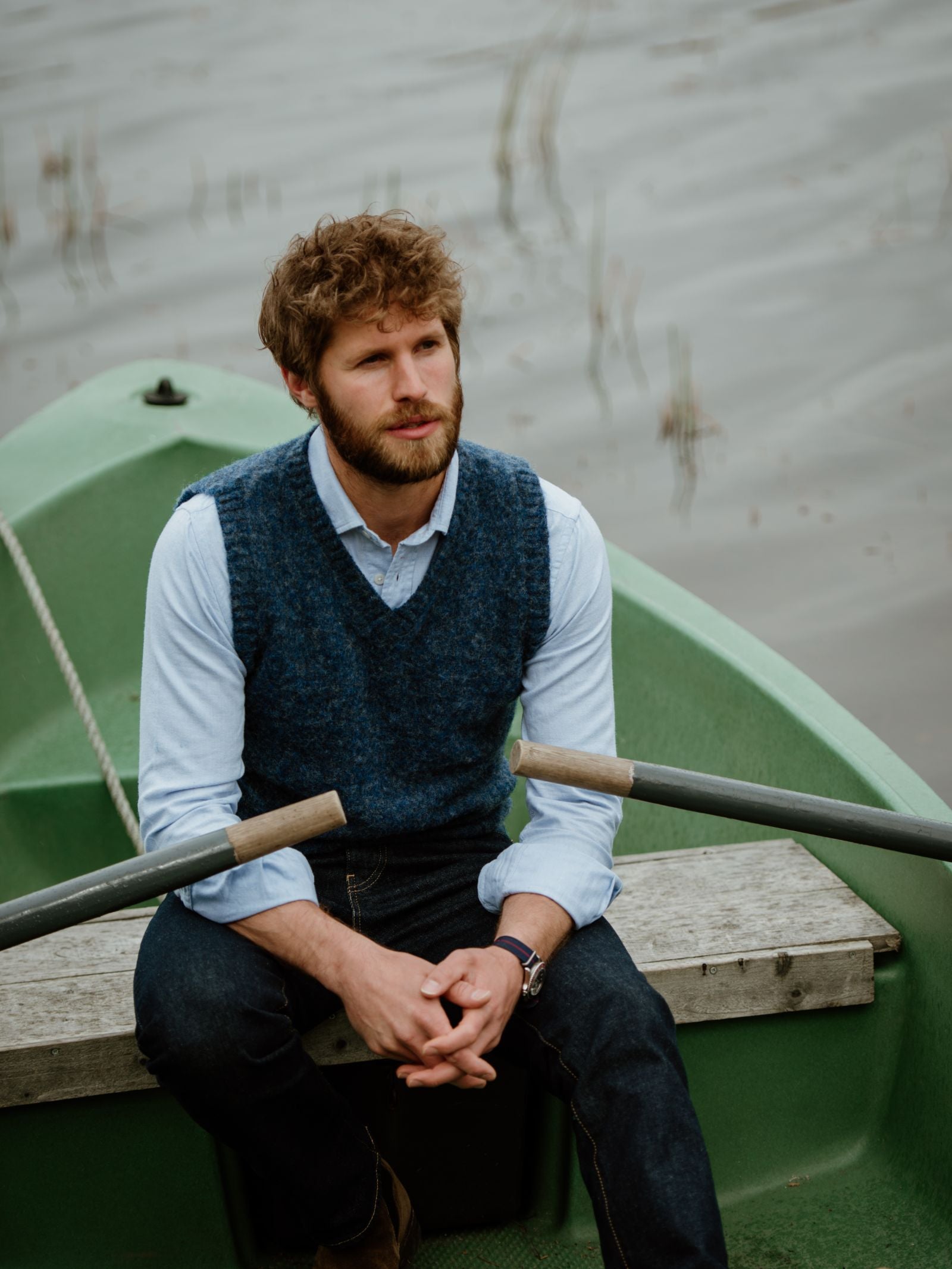 A bearded man with curly hair, dressed in Campbells of Beauly Shetland Slipover (a blue V-neck knit sweater vest) over a light shirt and jeans, sits thoughtfully in a green rowboat on calm water.
