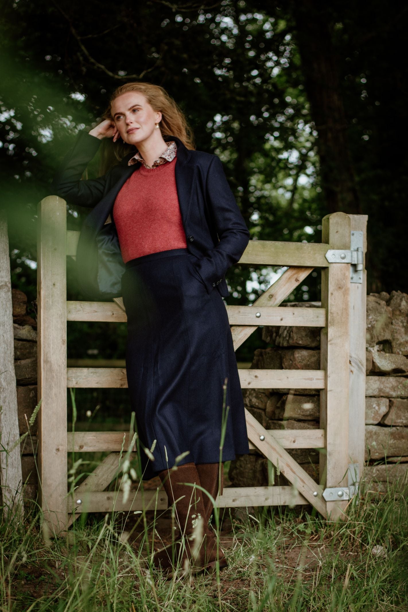 A woman with long, reddish hair stands outdoors by a wooden gate, wearing the Campbell's of Beauly Panel Skirt and coat over a red sweater. She looks thoughtful, one hand on her head, amid tall grass and trees.