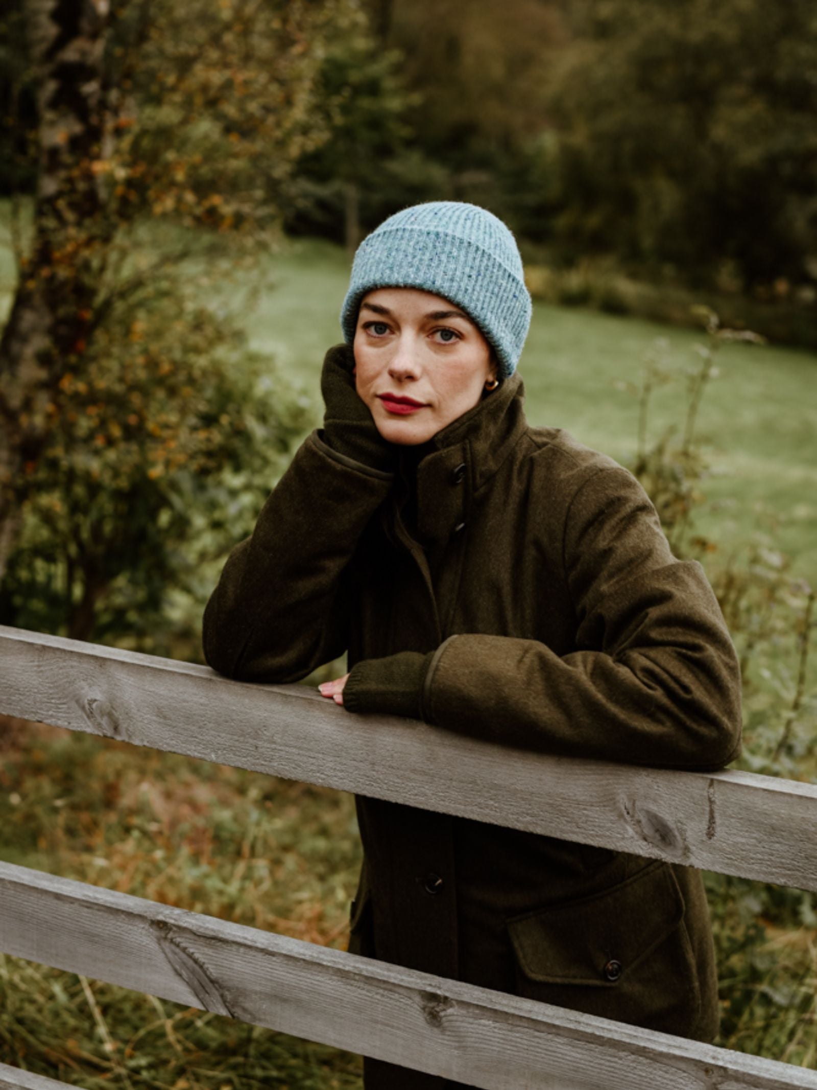 A woman in a blue Campbell's of Beauly Donegal Beanie and a dark green coat leans on a wooden fence, resting her chin on her hand, surrounded by grass and trees in the Scottish Borders.