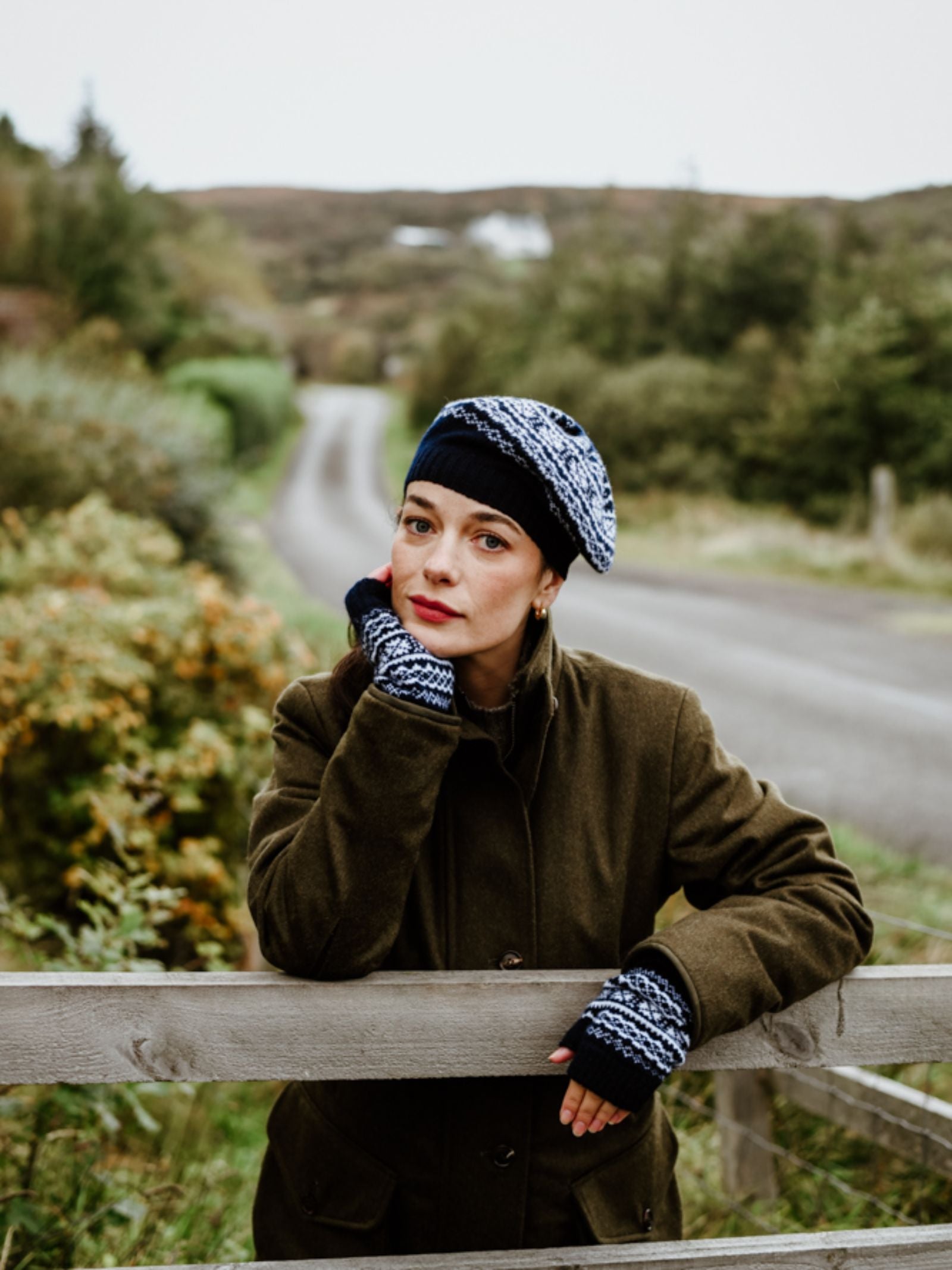 A woman in a brown coat, blue patterned hat, and Campbell's of Beauly Two-Colour Fairisle Half Finger Gloves leans on a wooden fence by a country road, with green trees and hills in the background.