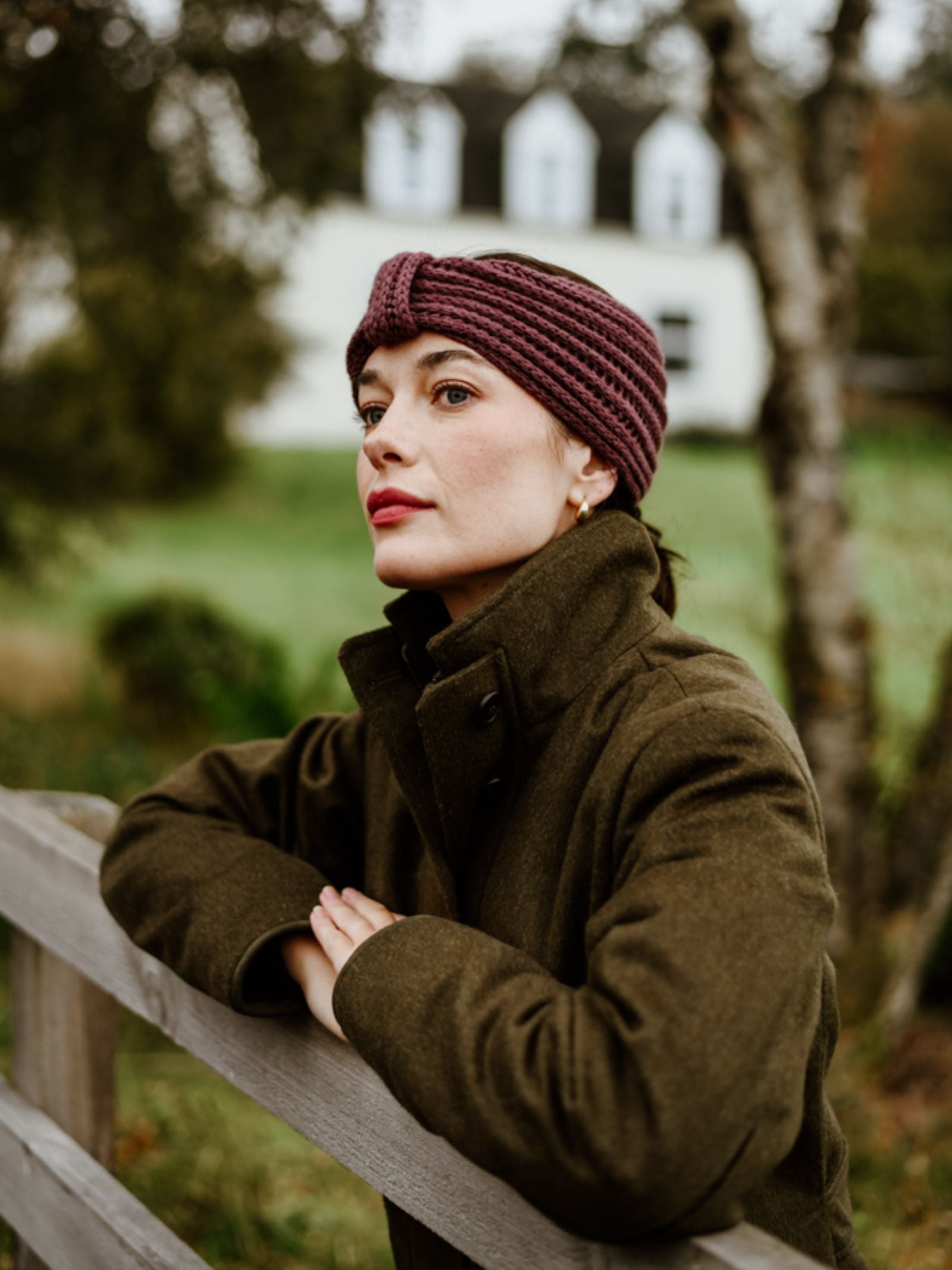 A woman in a brown coat wears the Campbell's of Beauly Cashmere Ear Warmer, leaning on a wooden fence outdoors with a blurred green yard and white house behind her—an elegant winter accessory scene.