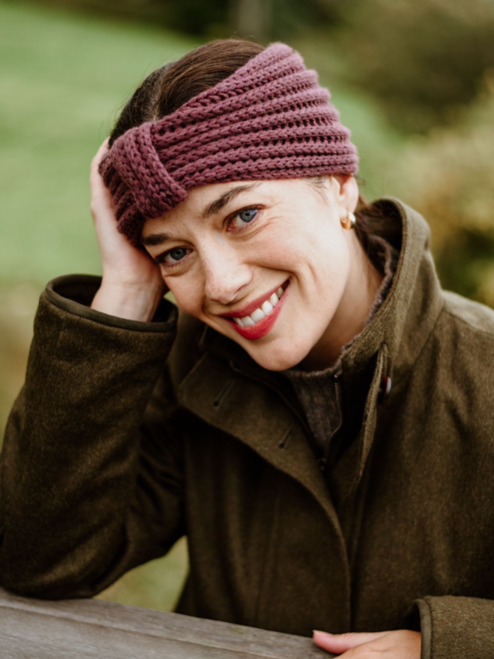 A woman wearing a Campbell's of Beauly Cashmere Ear Warmer in purple and an olive-green coat smiles warmly outdoors, resting her head on one hand against a grassy background, looking directly at the camera.