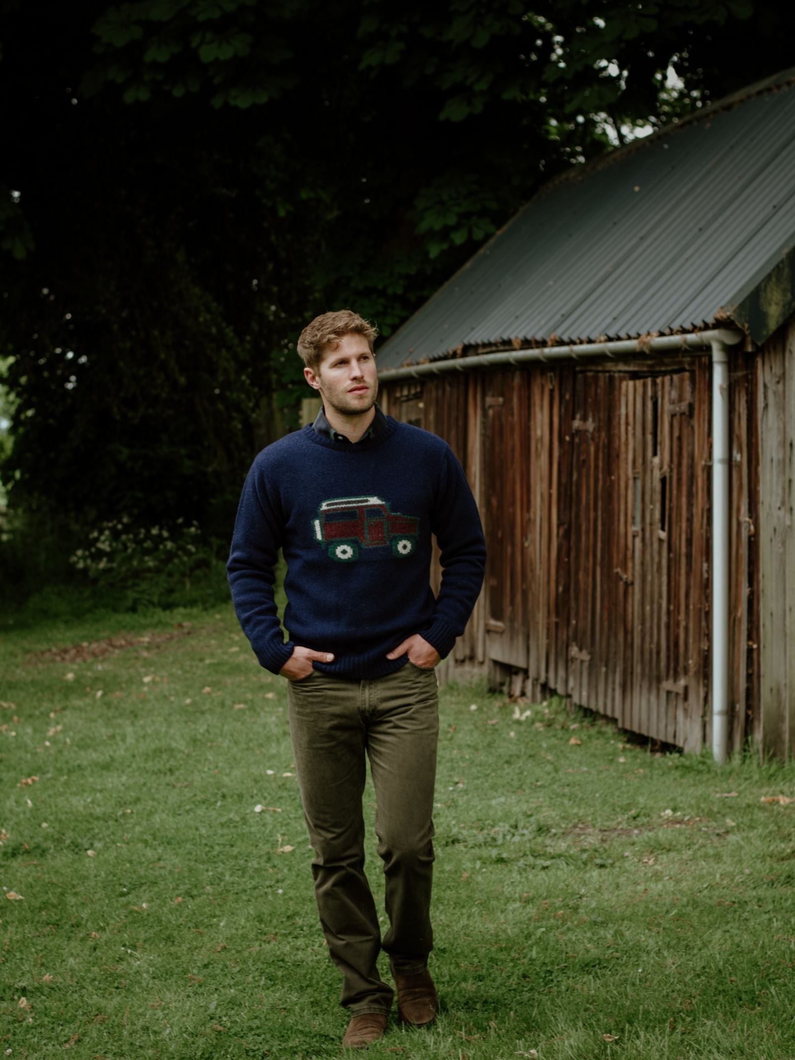 A man with short hair walks on grass near a rustic shed, wearing the Campbells of Beauly Land Rover Defender Jumper—a navy sweater with a red tractor motif—paired with olive pants, surrounded by trees and greenery.