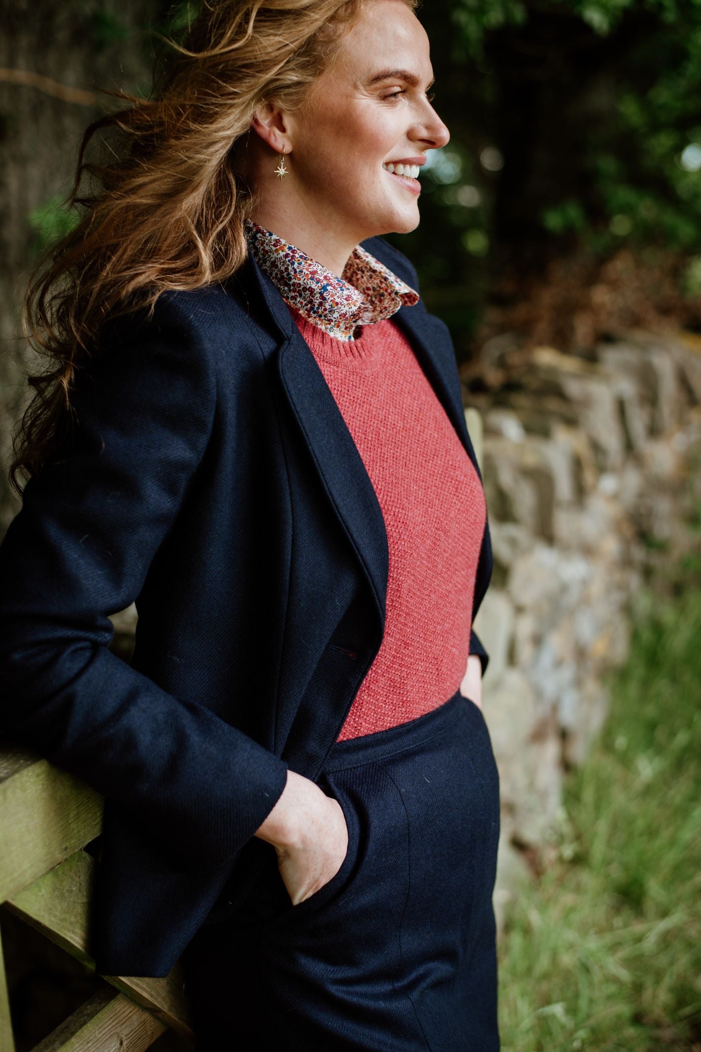 A person with long, light brown hair smiles outdoors by a stone wall, wearing a navy blazer, red sweater, floral-collared shirt, and the Panel Skirt by Campbell's of Beauly. Greenery surrounds them.