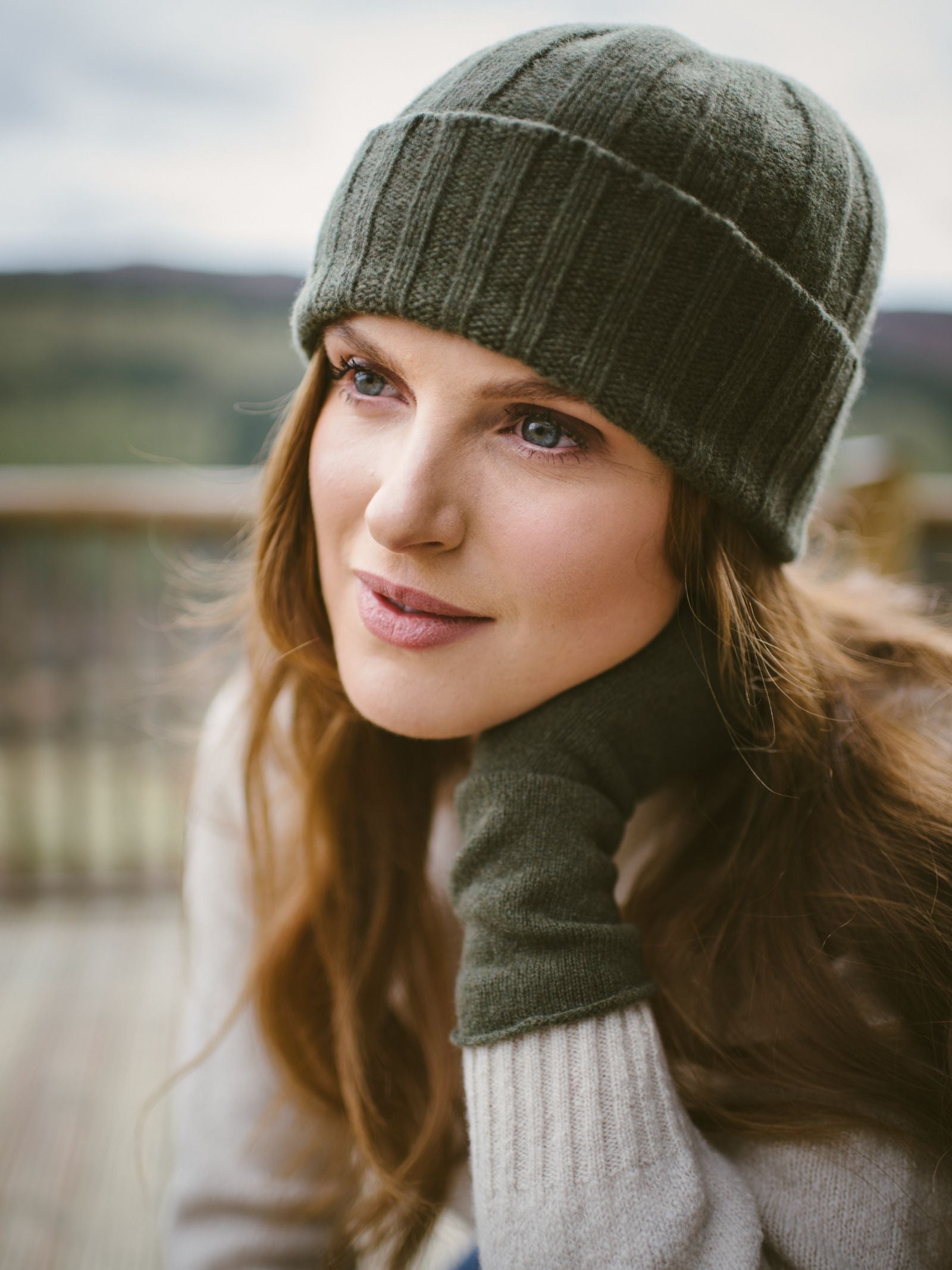 A woman with long brown hair wears a green knit hat and Campbells of Beauly Cashmere & Merino Fingerless Mittens, resting her chin on her gloved hand and gazing gently to the side, with a softly blurred outdoor background behind her.