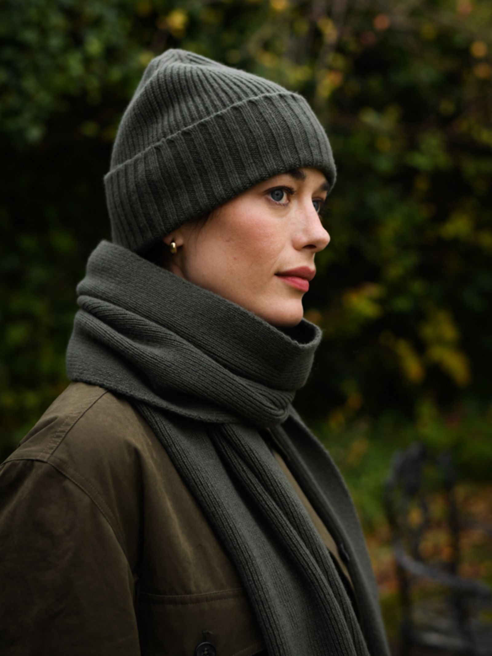 A woman wears the Campbell's of Beauly Rib Hat in green, paired with a matching UK-knit scarf and olive coat. She stands outdoors amid lush greenery—a true winter essential.