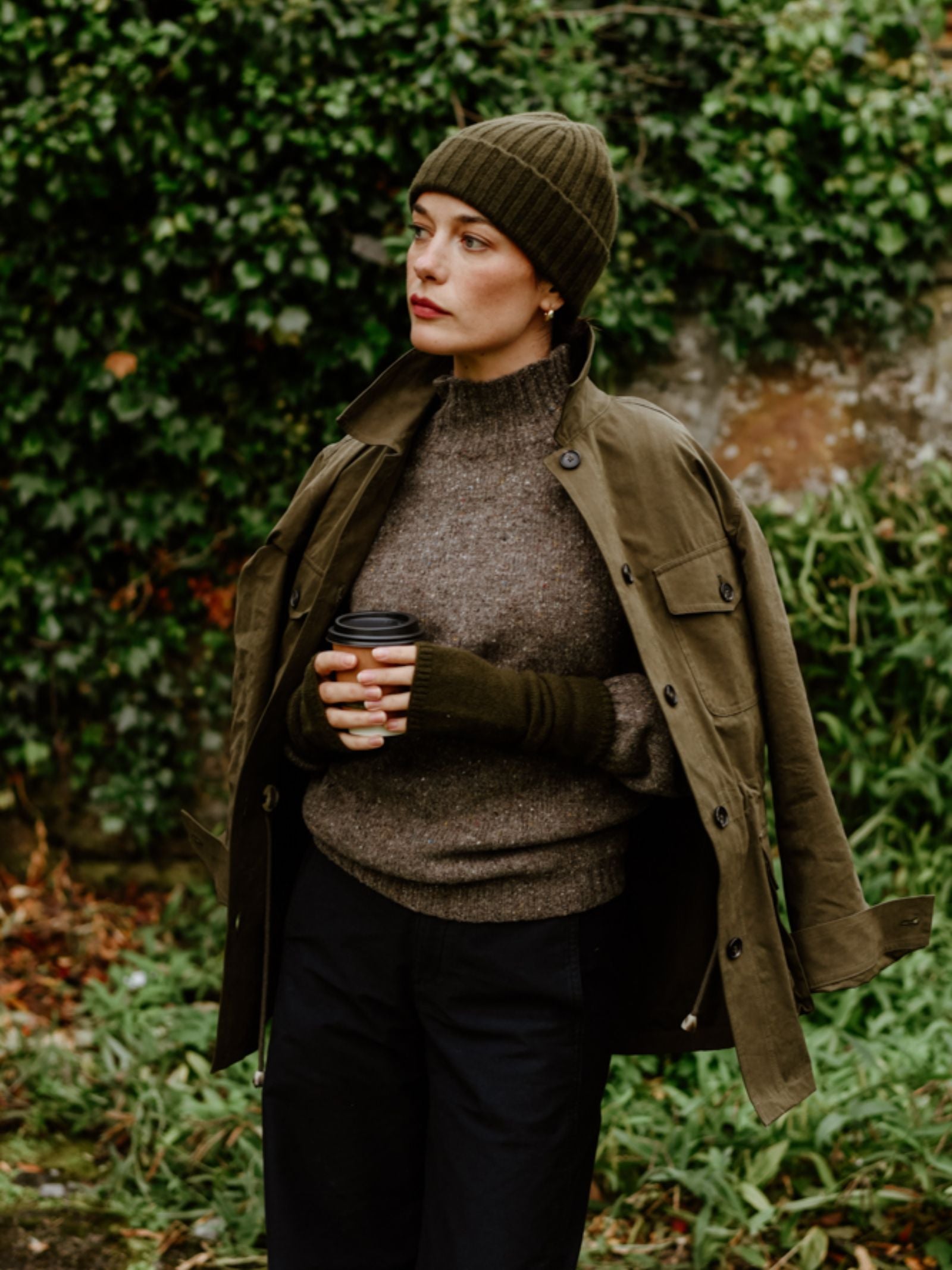 A woman stands outdoors against green ivy, wearing a Campbell's of Beauly Cashmere Rib Hat, an olive jacket over her shoulders, a brown knit sweater, and dark pants, holding a takeaway coffee cup in both hands.