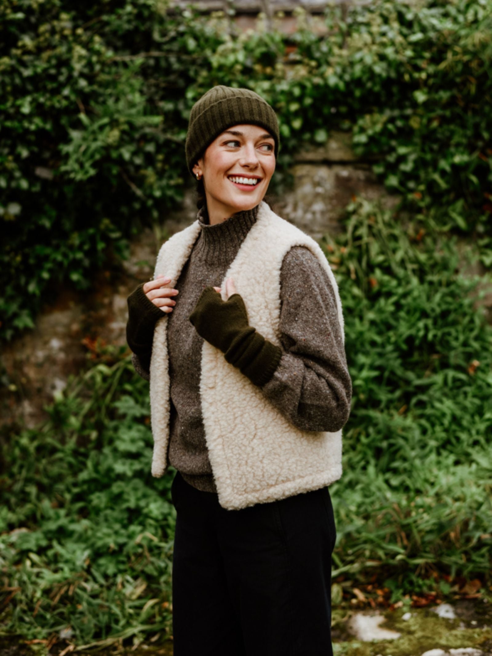 A smiling woman stands outdoors before green foliage, wearing the Campbell's of Beauly Donegal Lambswool Funnel Neck Jumper, a green knit beanie, cream sherpa vest, fingerless gloves, and black pants. She looks to the side with hands near her vest.