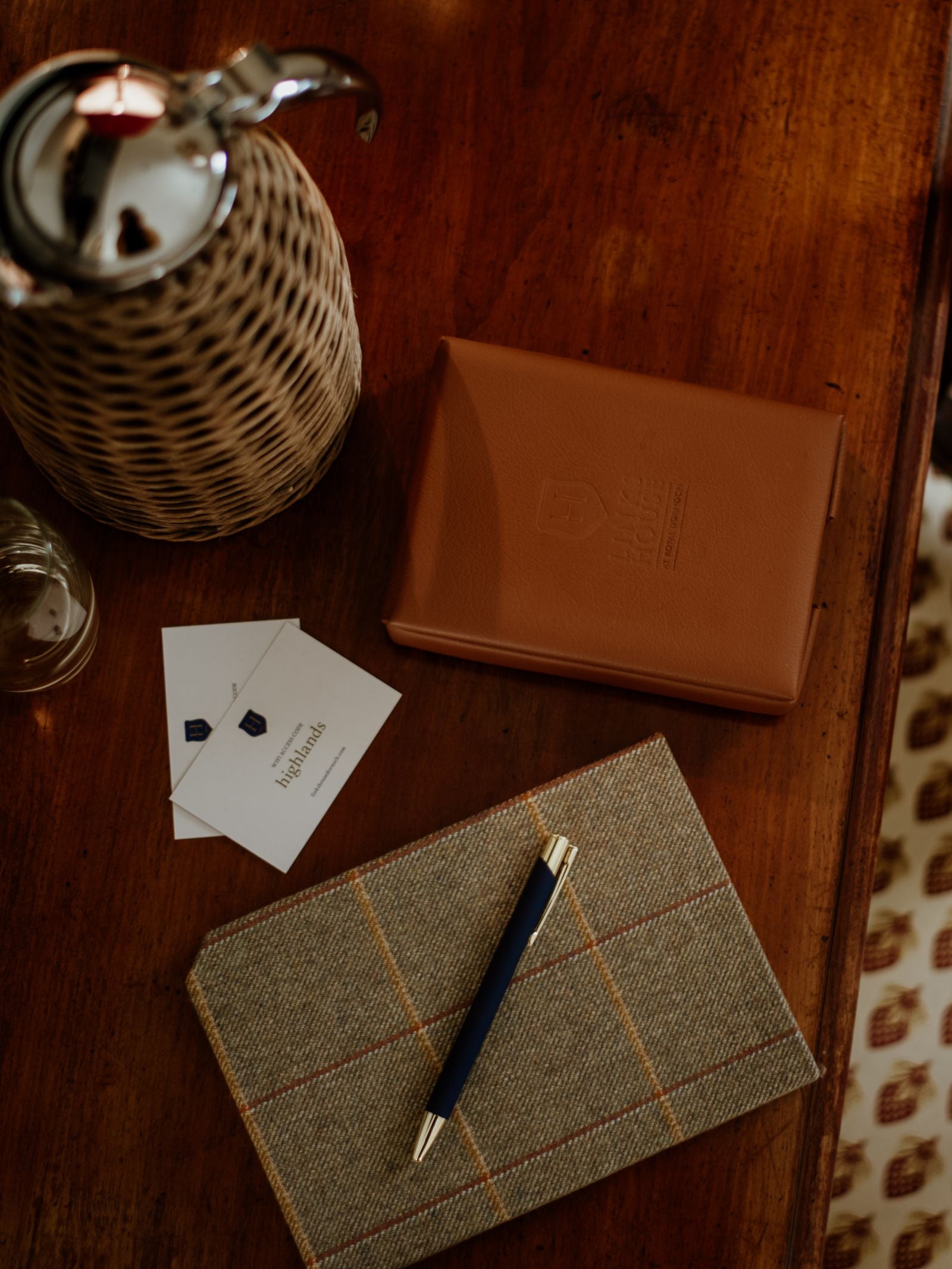 A Campbells of Beauly Tweed Notebook sits atop a wooden desk alongside a pen, brown leather folder, business cards, and a wicker-covered pitcher.