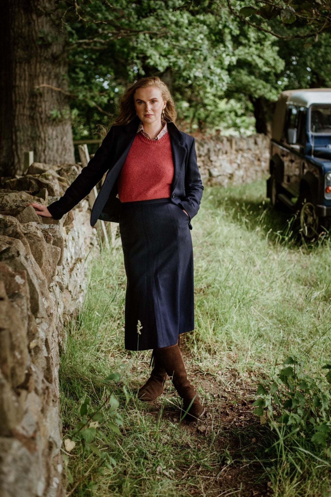A woman stands outdoors by a stone wall, wearing Campbell's of Beauly Panel Skirt with a navy blazer, red sweater, and brown boots. She is on grass near trees, with a vintage off-road vehicle in the background.