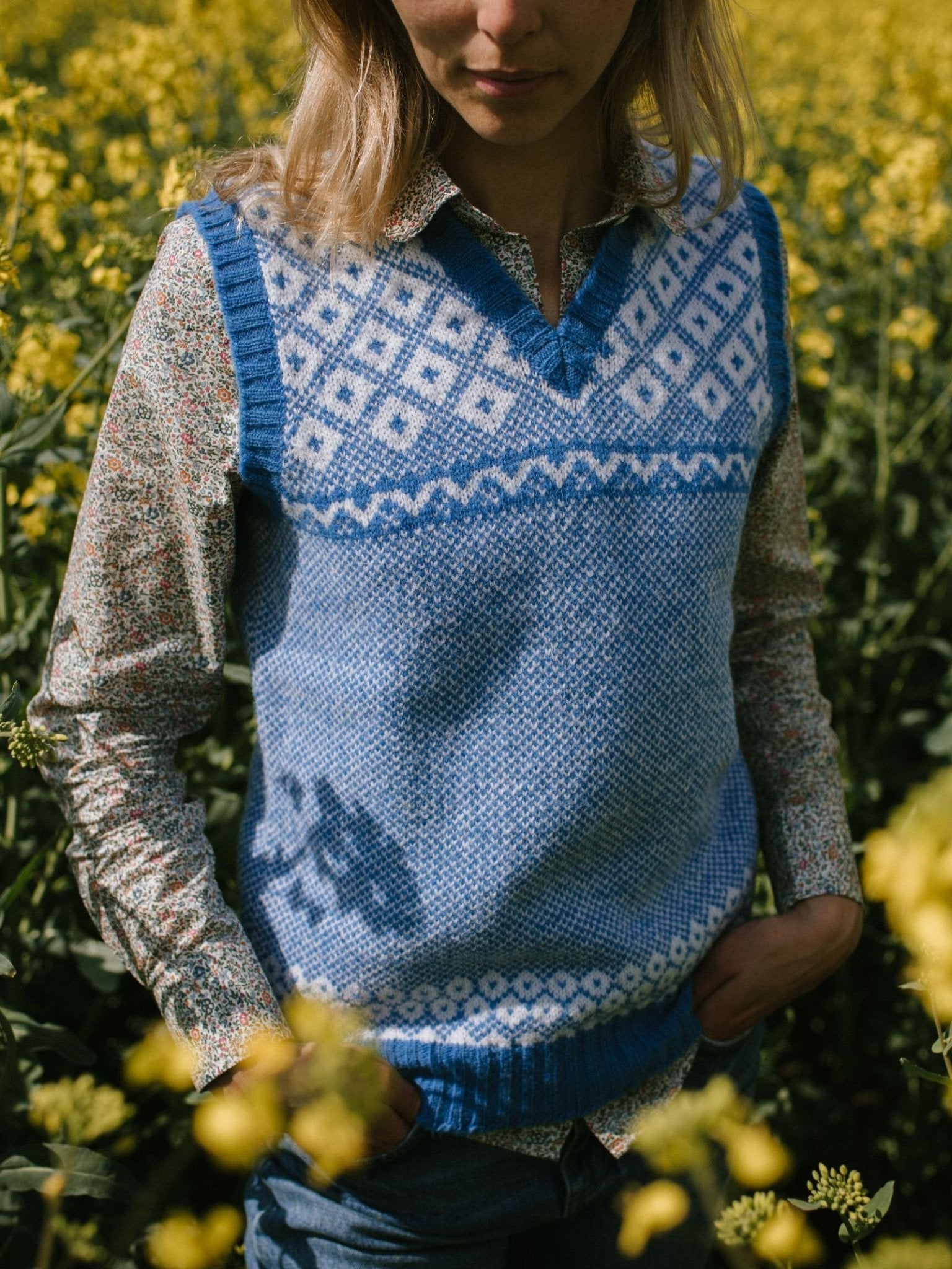 A person stands in a field of yellow flowers wearing the Campbells of Beauly Nordic V Tank over a floral shirt, hands in jeans pockets. Their face is partially out of frame.