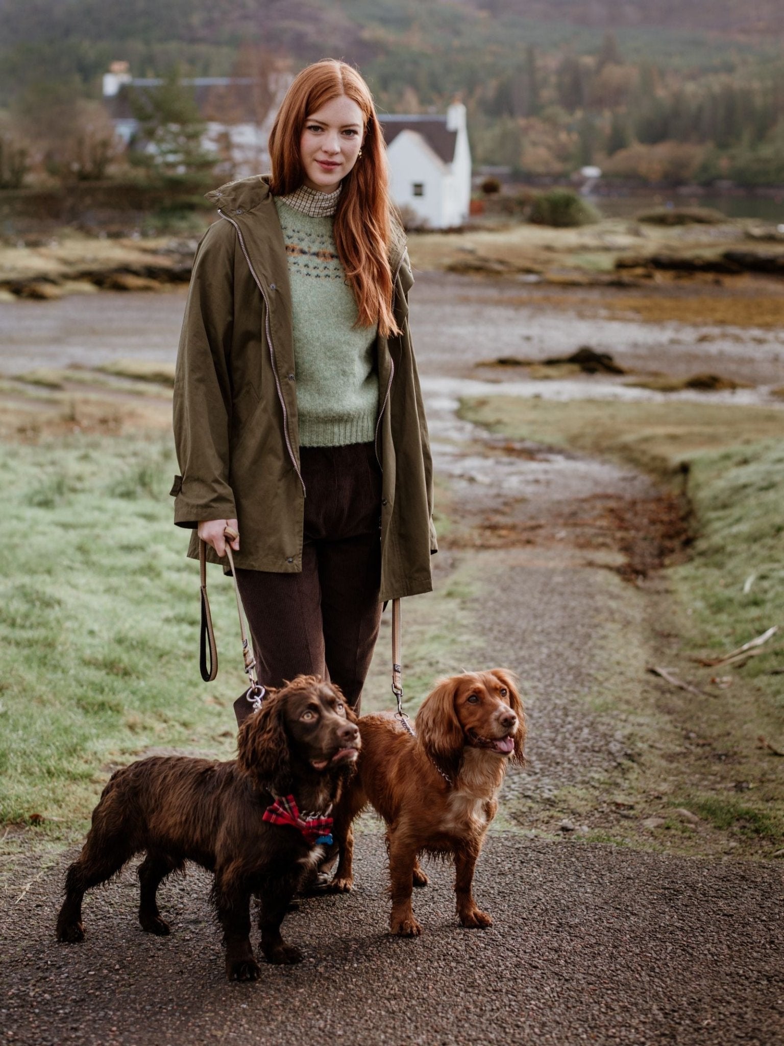 A woman with long red hair wears the Auld Stock Cropped Fairisle Crew Neck Jumper by Campbell's of Beauly as she stands outside on a path, holding leashes and walking two small brown dogs, with a white house and trees in the background.