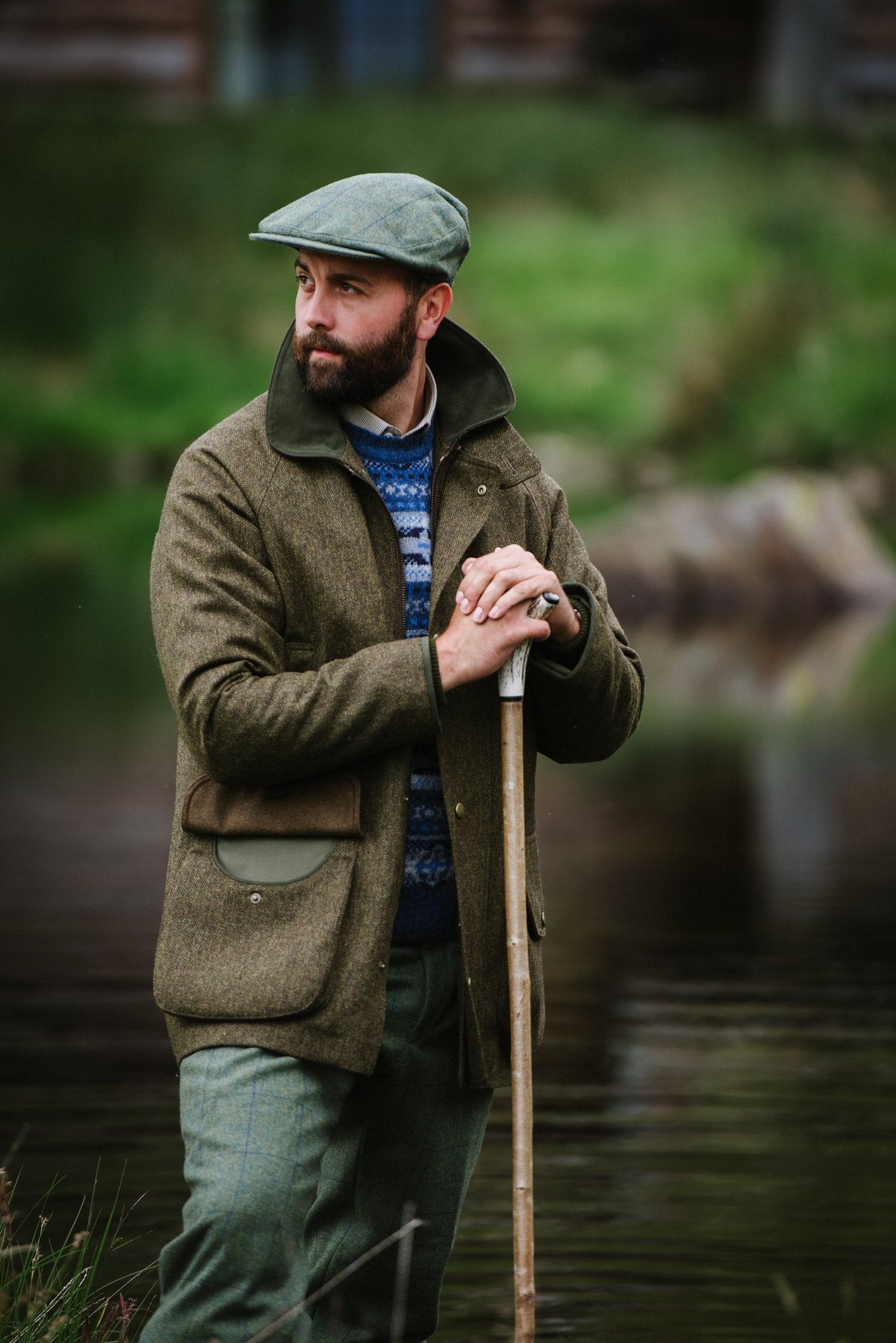 A bearded man in a flat cap and Campbells of Beauly Tweed Field Coat with an Alcantara collar stands by a calm river, holding a wooden walking stick, surrounded by greenery and rocks.