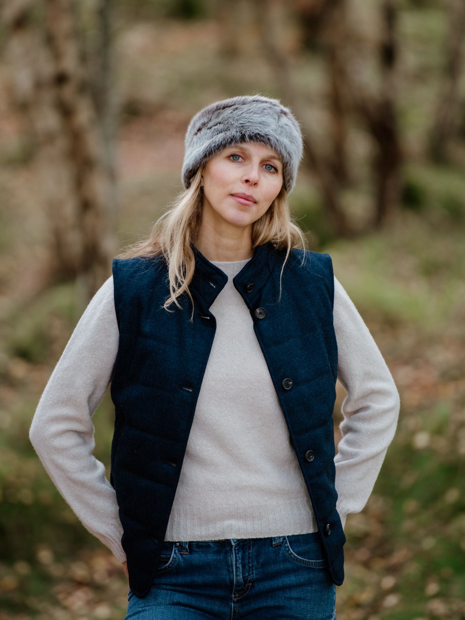 A woman stands in a forest wearing a gray Sheepskin Headband by Campbells of Beauly, a navy blue buttoned vest over a light sweater, and blue jeans. She has blonde hair and looks at the camera with a neutral expression.