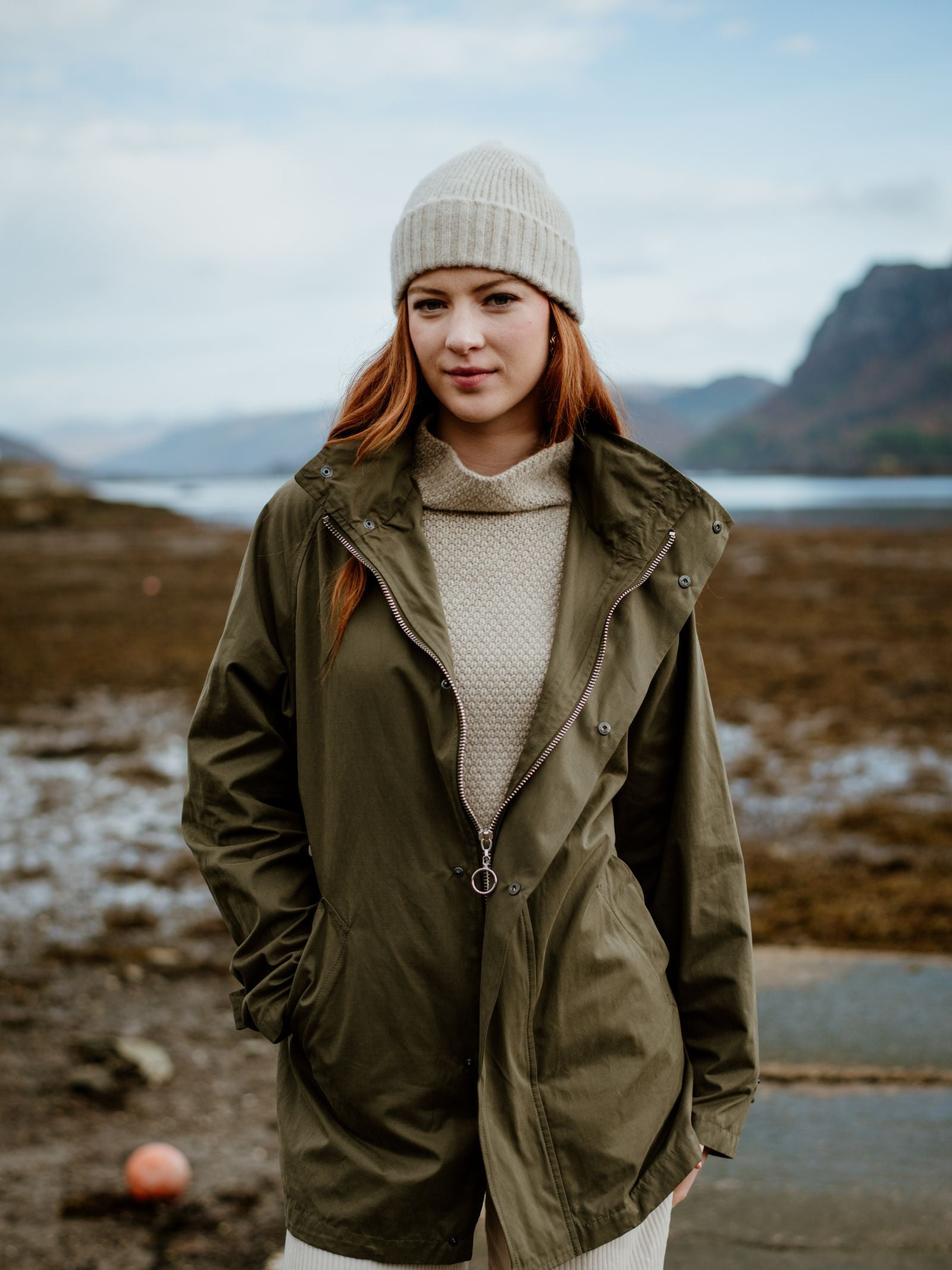 A young woman with long auburn hair wears a Campbells of Beauly Lambswool Hat, a cream sweater, and an olive green jacket as she stands outdoors by a rocky shoreline with mountains and water in the background.