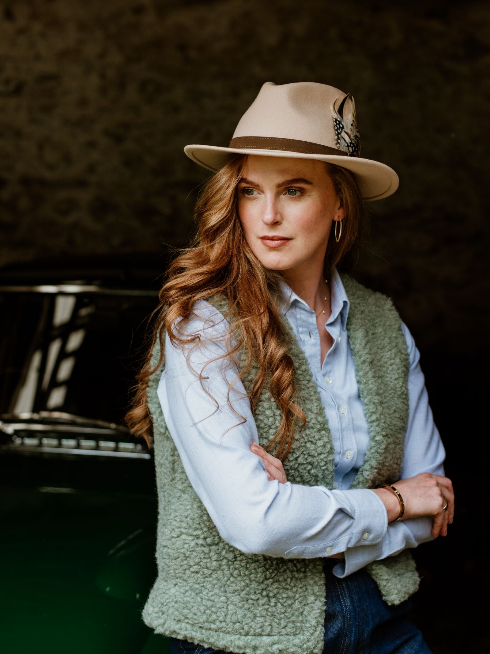 A woman with long wavy hair, wearing a Hicks & Brown Gamebird Fedora with a feather, light blue shirt, and green vest stands with arms crossed in front of a dark background and a classic car.