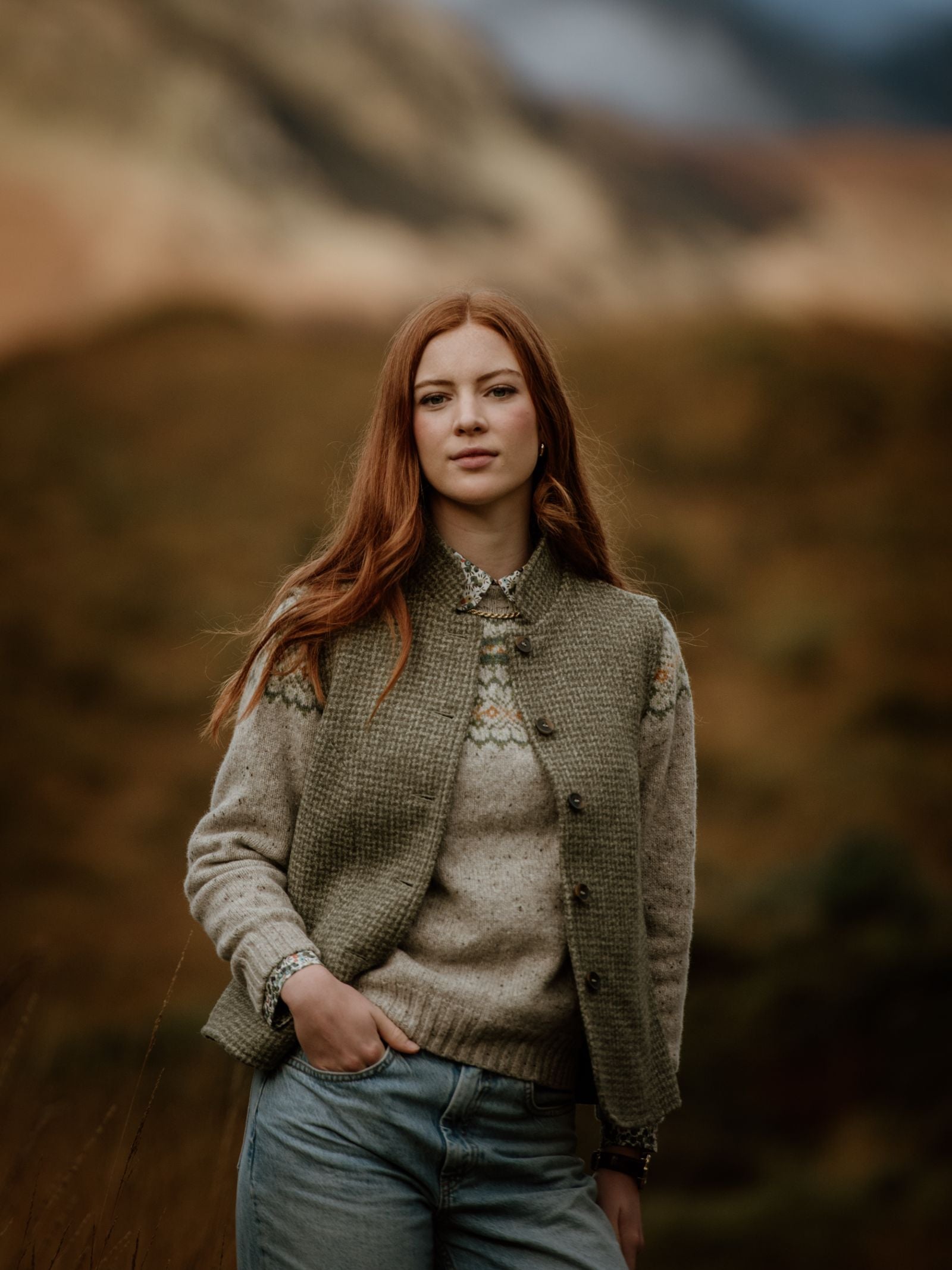 A young woman with long red hair stands outdoors before a blurred natural backdrop, wearing jeans, a knit sweater, a patterned shirt, and the Campbell’s of Beauly Harris Tweed Houndstooth Gilet, looking calmly at the camera.