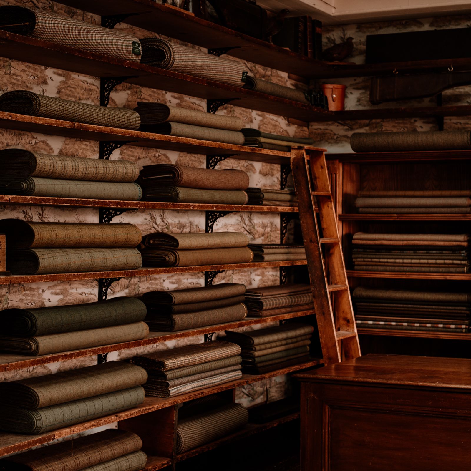 Bolts of patterned fabric in various earthy tones are neatly stacked on wooden shelves at Campbells of Beauly, set against a rustic wall with a wooden ladder nearby and part of a wooden counter visible in the foreground.