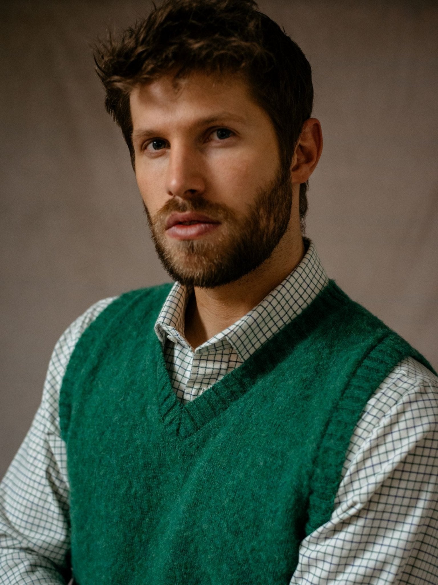 A man with short brown hair and a beard models the Campbells of Beauly Shetland Slipover—a green V-neck knit sweater vest—over a checked shirt, looking slightly to the side against a neutral background.