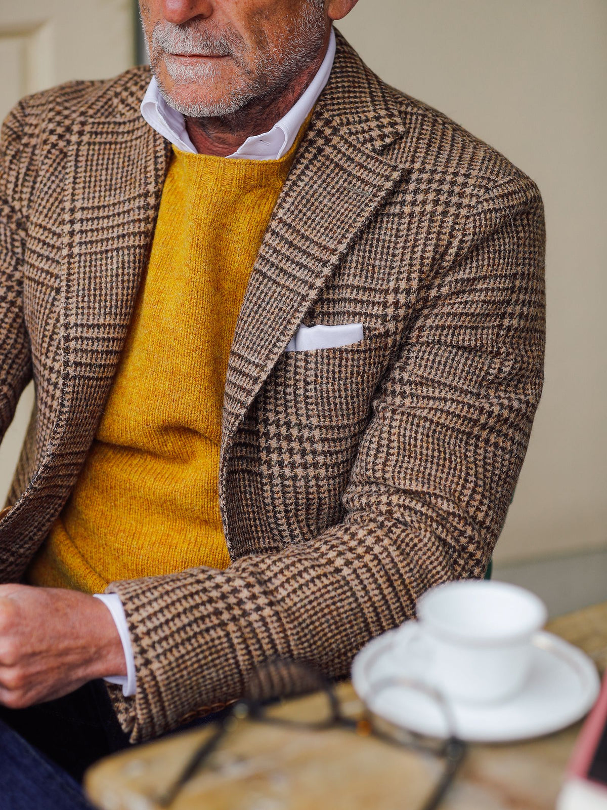 A man wearing a Campbell’s of Beauly Harris Tweed Jacket with Italian tailored flair, paired with a yellow sweater and white shirt, sits at a table with a white cup and saucer. His face is partially visible, and a white pocket square accents his blazer.