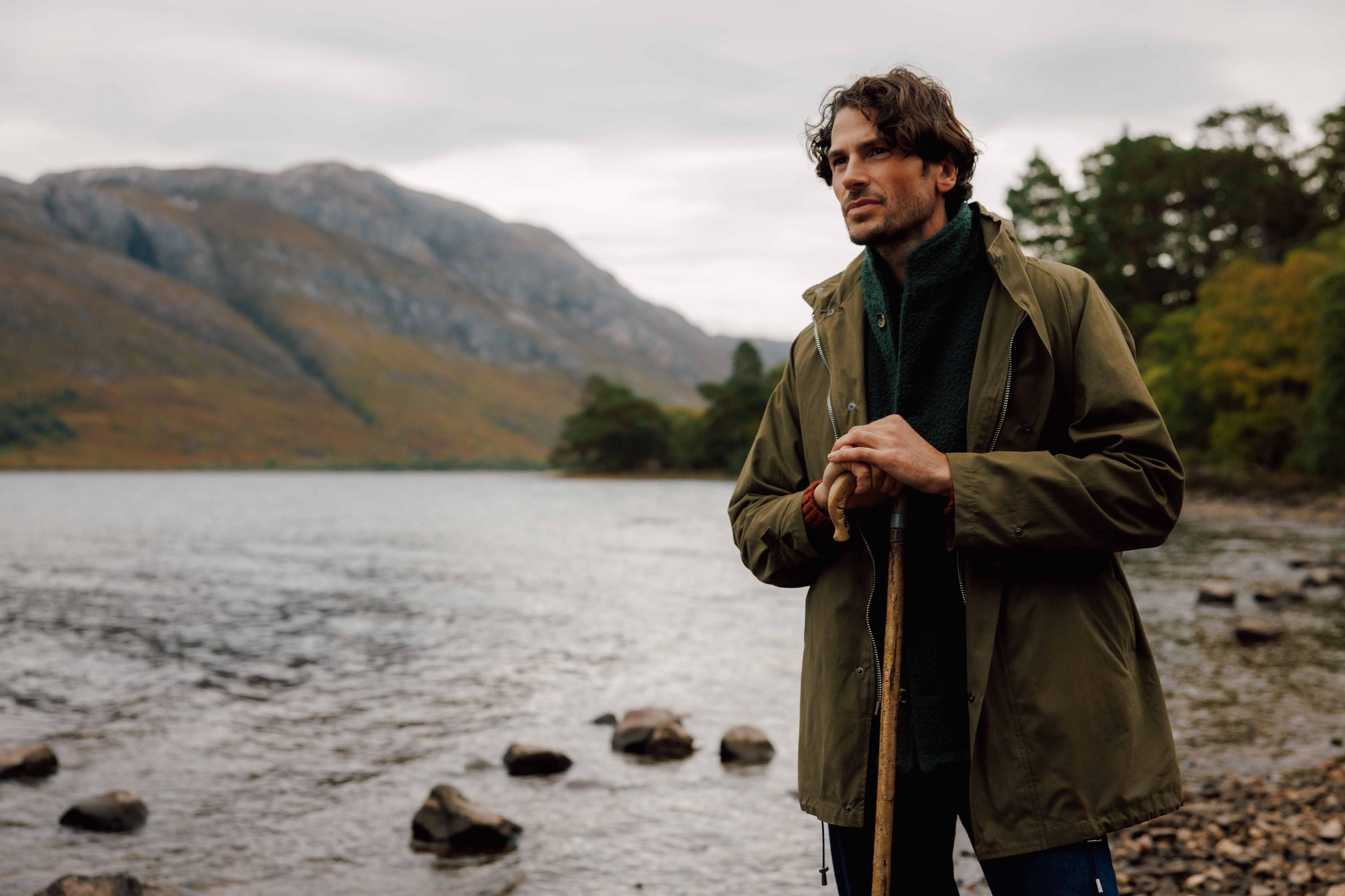A man in a green jacket stands on a rocky lakeshore, holding a wooden walking stick, with mountains and trees in the background under a cloudy sky.
