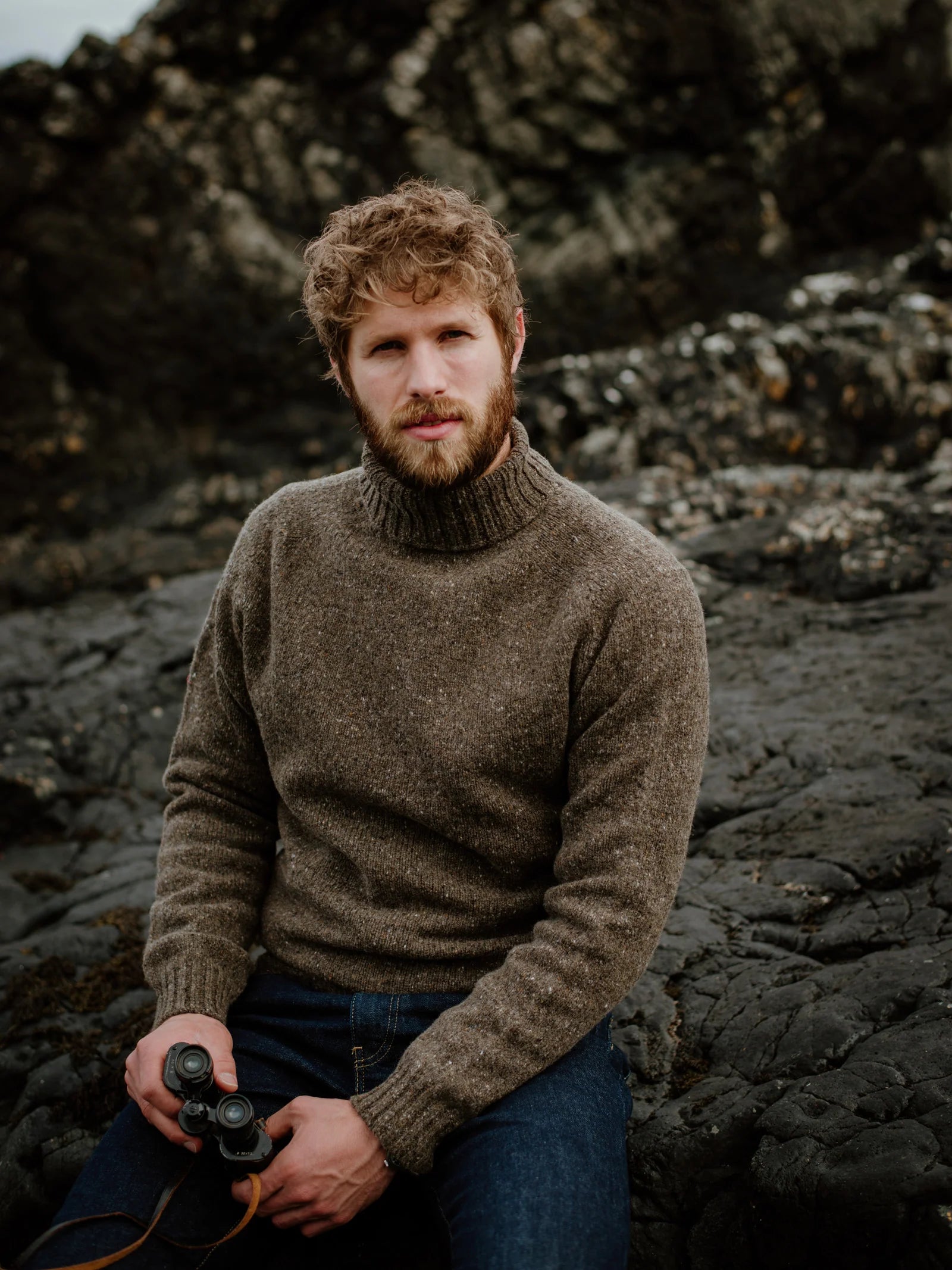 A man with curly hair and a beard sits on dark rocky terrain, wearing jeans and the Campbell’s of Beauly Donegal Lambswool Poloneck, holding binoculars and looking toward the camera.