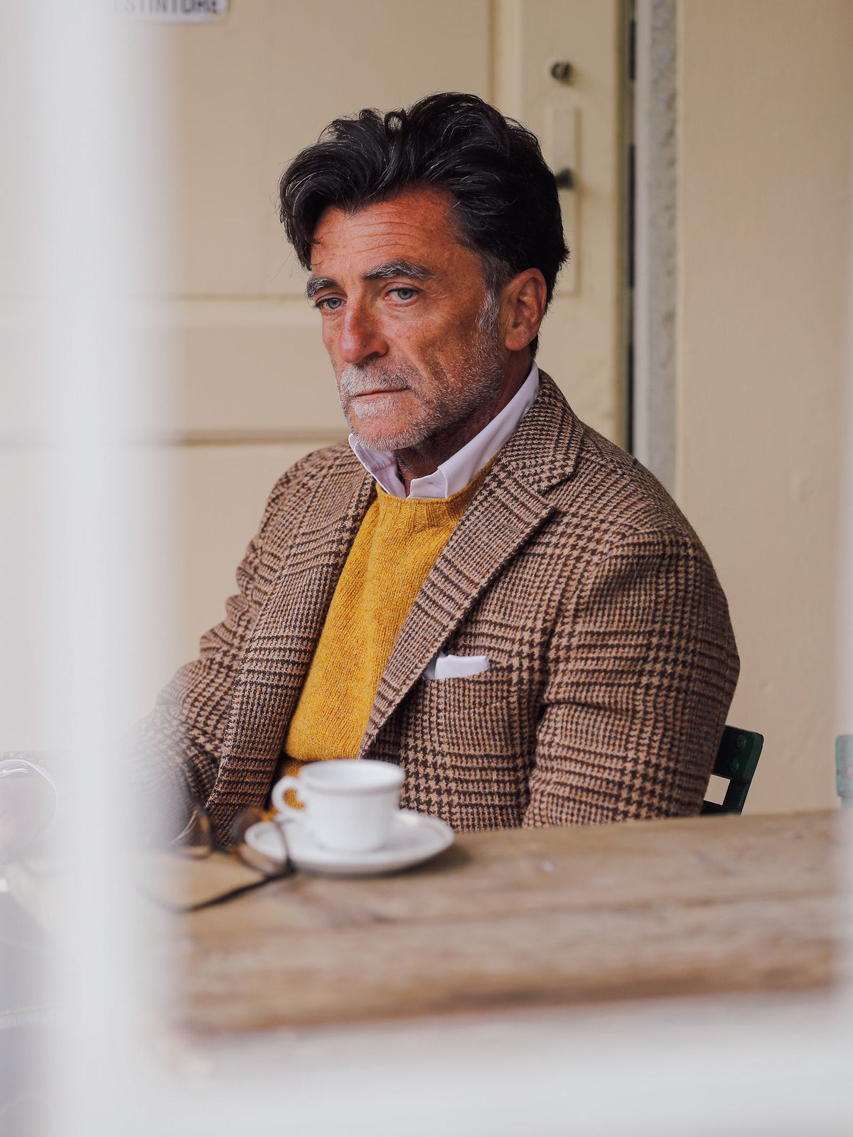 A man with dark hair and a beard, dressed in the Campbell’s of Beauly Harris Tweed Jacket in brown plaid, sits thoughtfully at a table with a white coffee cup before him, a yellow sweater layered beneath his jacket.