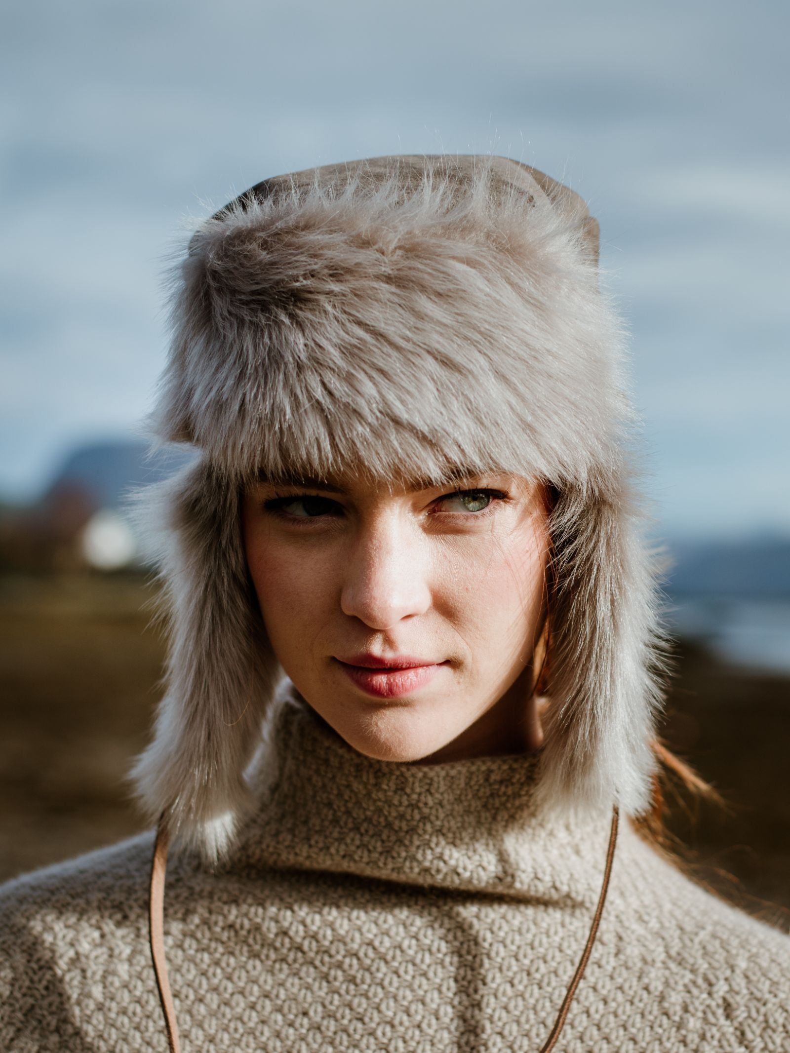 A woman stands outdoors in a Campbells of Beauly Toscana Sheepskin Trapper Hat, its light-gray, fluffy ear flaps paired with a beige knit turtleneck sweater. She gazes to the side, exuding cold-weather comfort against a blurred natural backdrop.