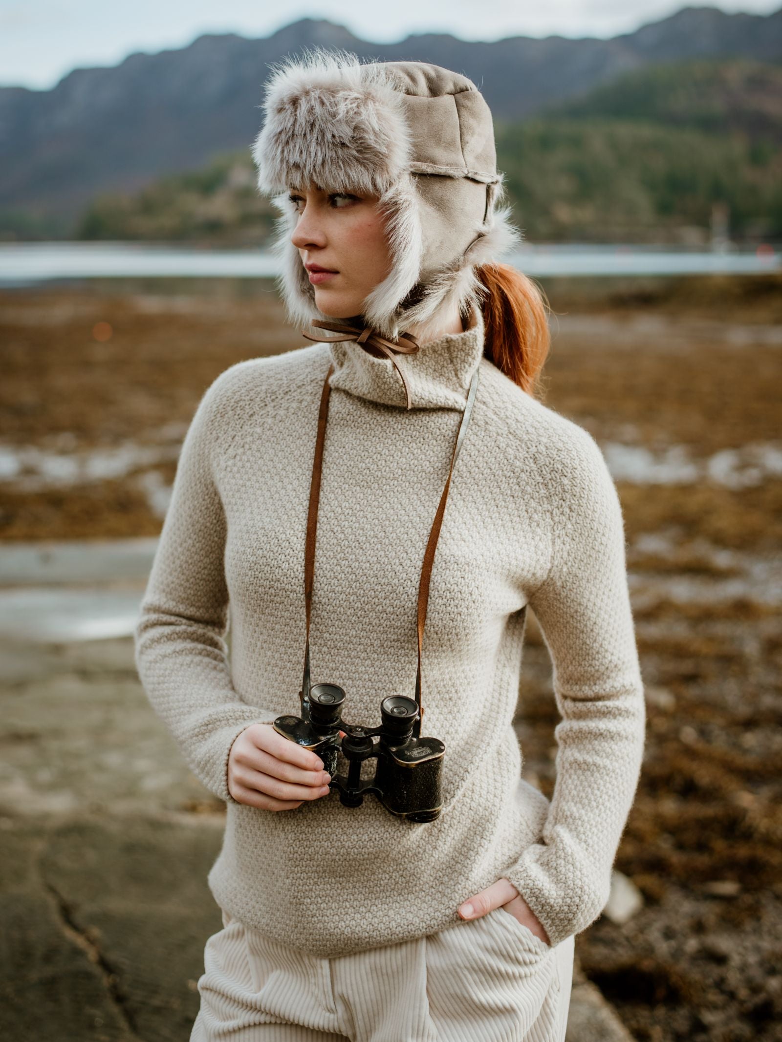 A woman stands on a rocky shore, wrapped in a beige textured sweater and light pants. She wears the Campbells of Beauly Toscana Sheepskin Trapper Hat with ear flaps and gazes into the distance, binoculars hanging from her neck.