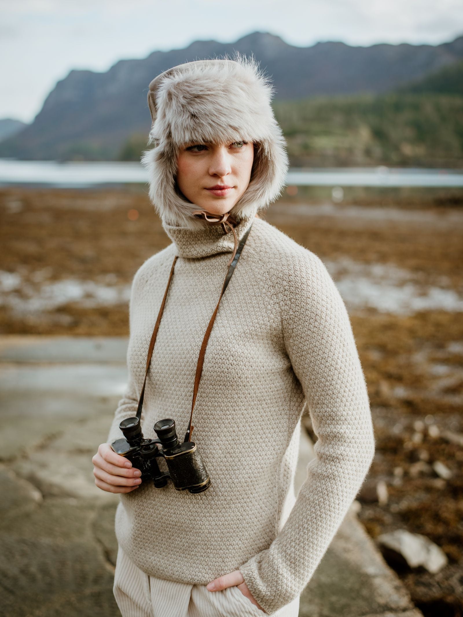 Wearing the Campbells of Beauly Toscana Sheepskin Trapper Hat and a textured beige sweater, a person stands by water and mountains, binoculars in hand, embodying cold-weather comfort as they gaze to the side.