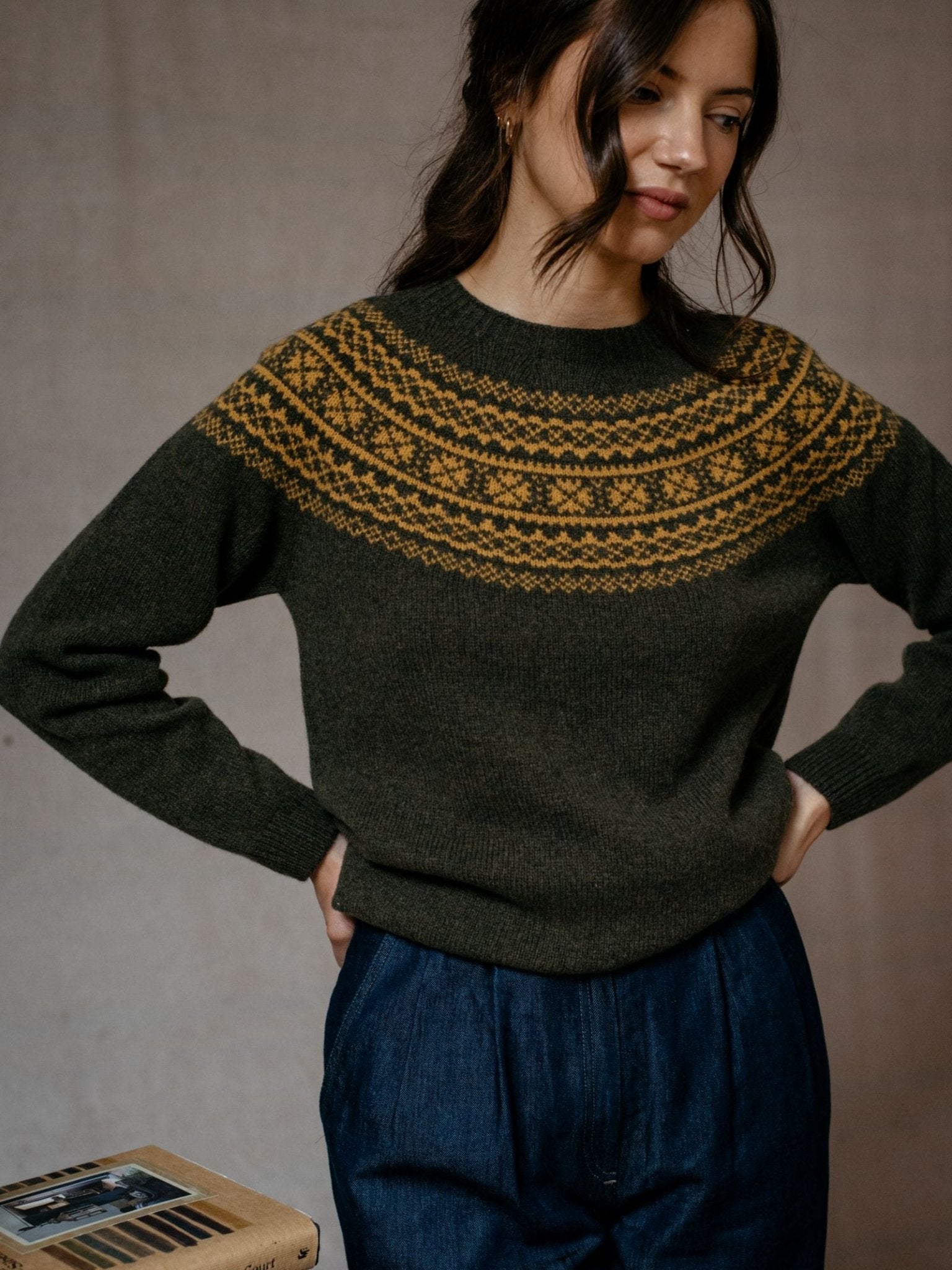 A woman stands with her hands on her hips, wearing the Campbells of Beauly Two-Colour Fairisle Yoke Jumper, which features a dark green base and yellow geometric pattern, next to a small table with a photo album.