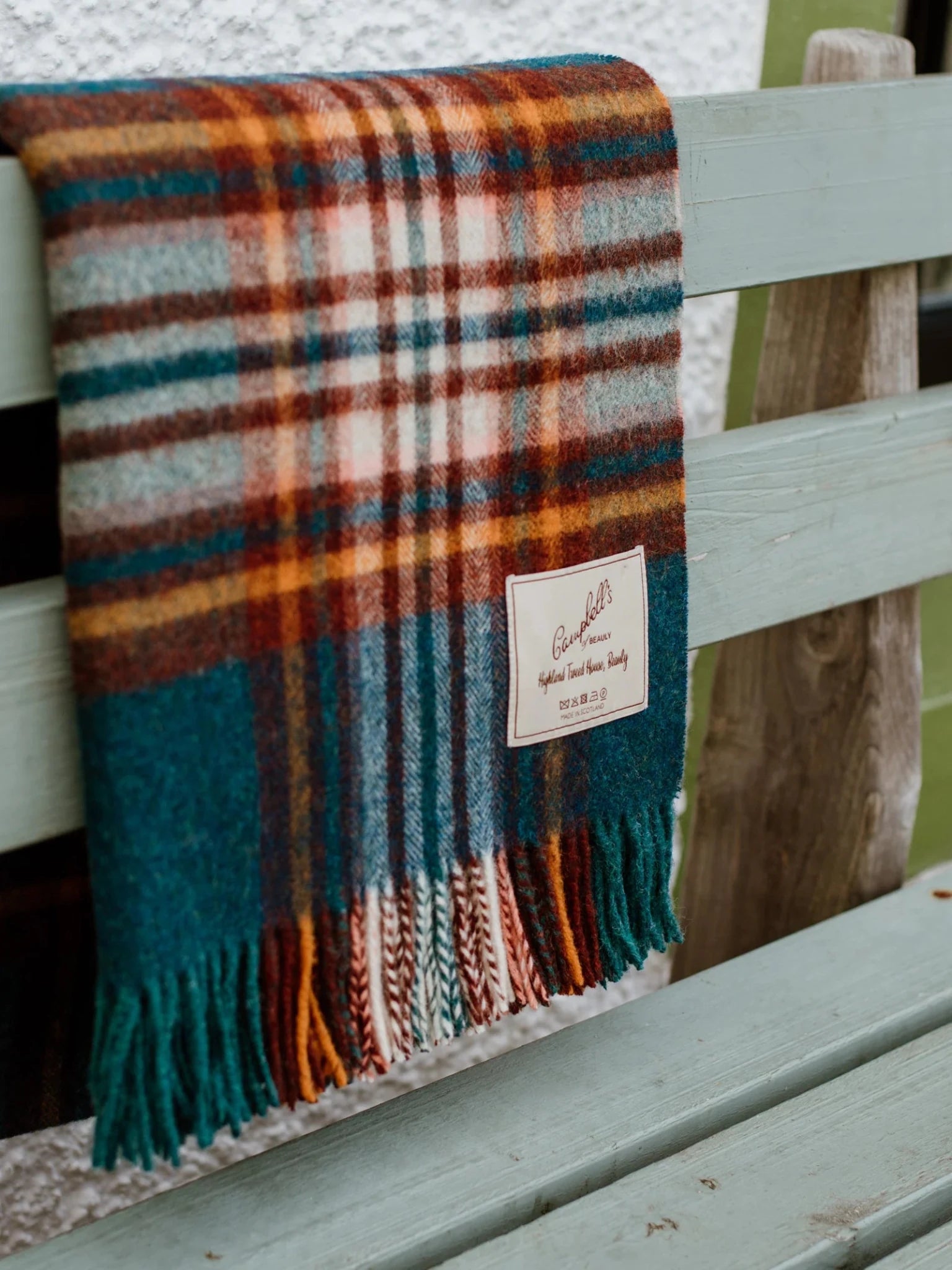 The Auld Stock Abriachan Lambswool Throw by Campbell's of Beauly, with fringe and label, adds a splash of color draped over a weathered light green wooden bench outdoors.