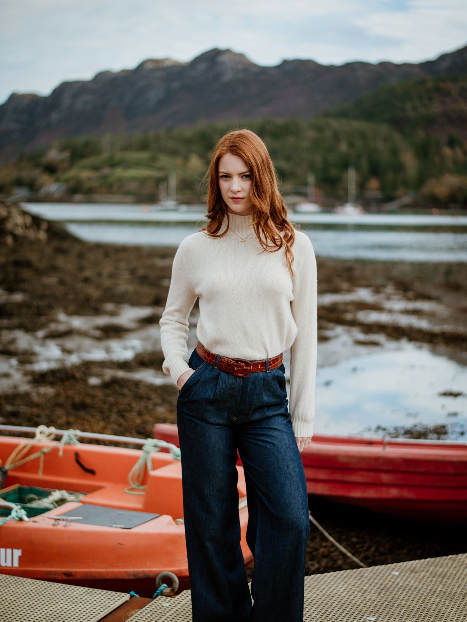 A woman with long red hair, wearing the Campbells of Beauly Geelong Lambswool Funnel Neck sweater and wide-leg jeans, stands on a dock by orange boats, with mountains and water in the background.